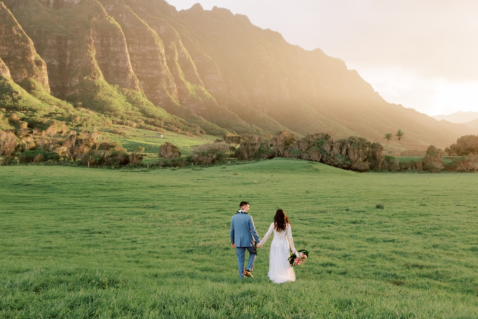 A bride and groom walking through a grassy field with the view of the mountains at sunset.