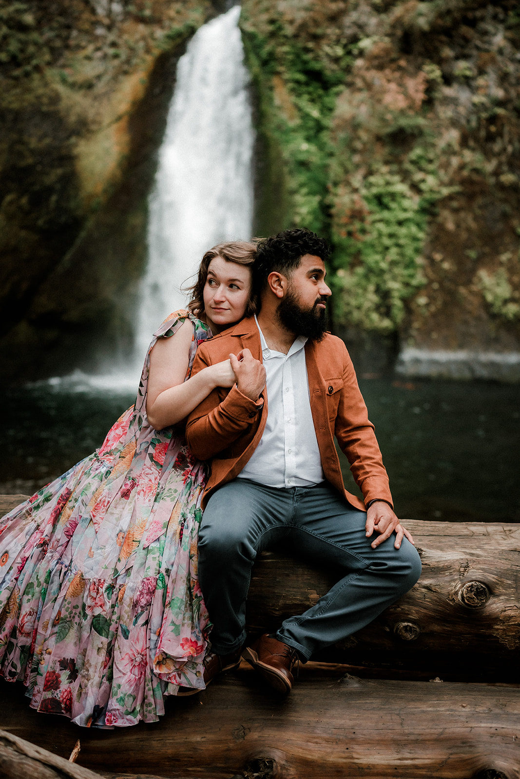 couple sitting on log pile in front of Wahclella Falls for their engagement photo session in Portland, OR
