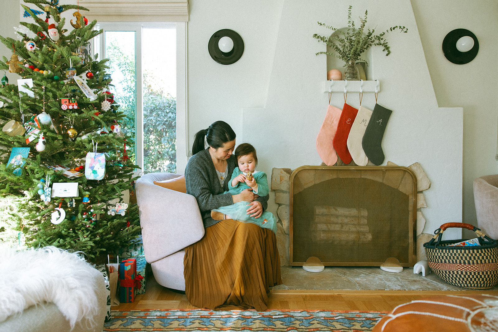 asian mom and baby sitting in living room with christmas tree and stocking hanging over the fireplace