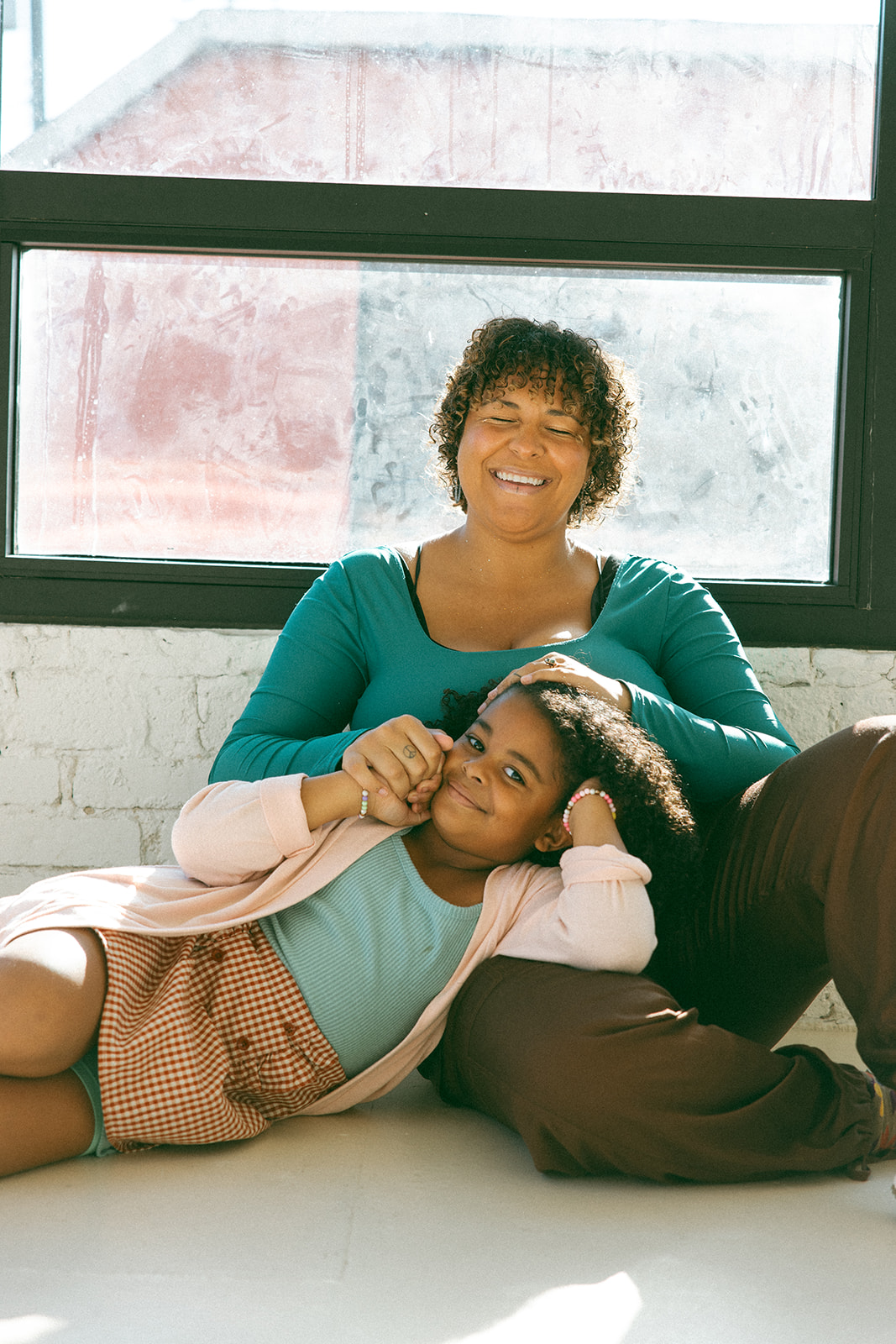 black mom sitting on floor in front of large window with young daughter leaning into her lap