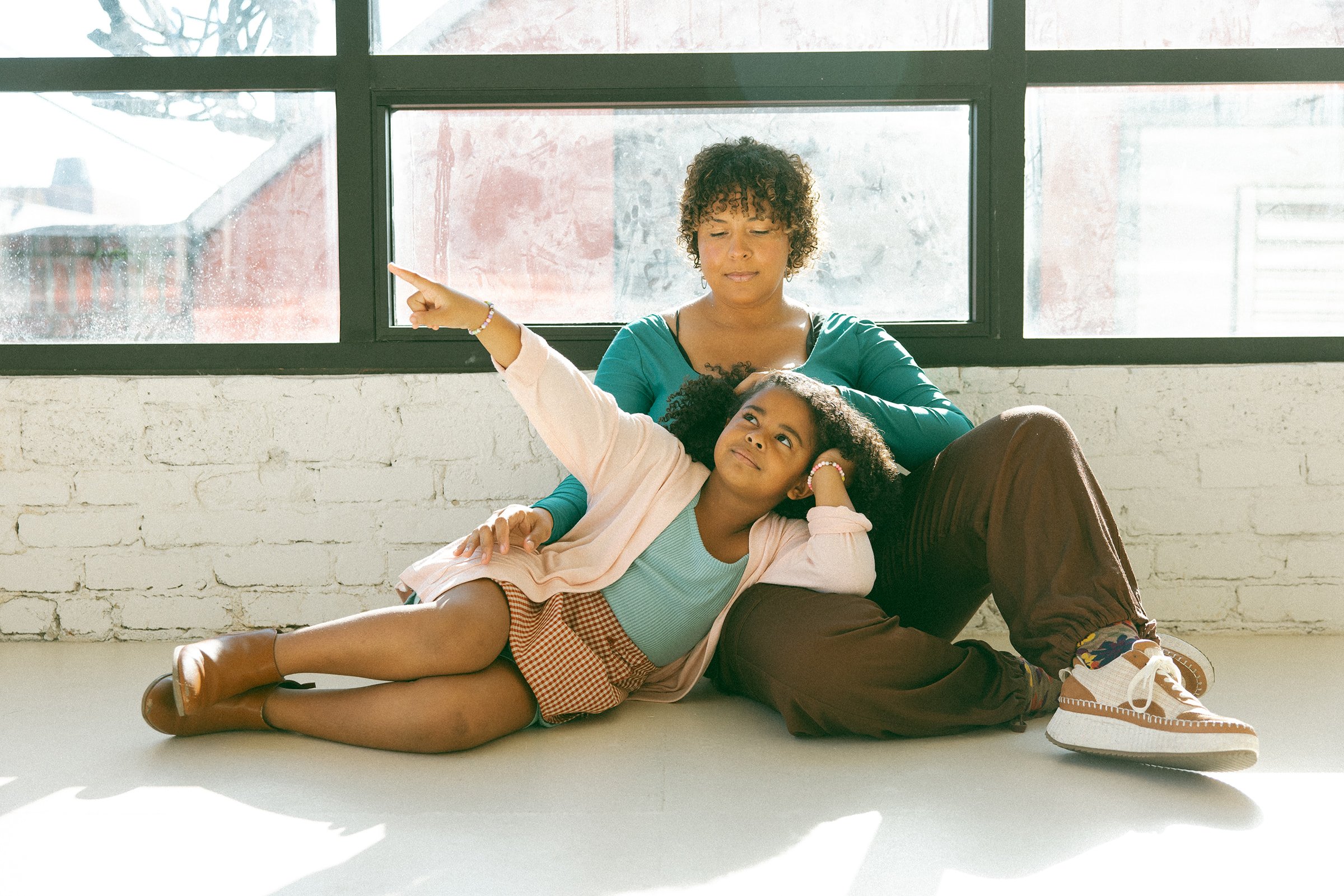 black mom sitting on floor in front of large window with young daughter laying across her lap