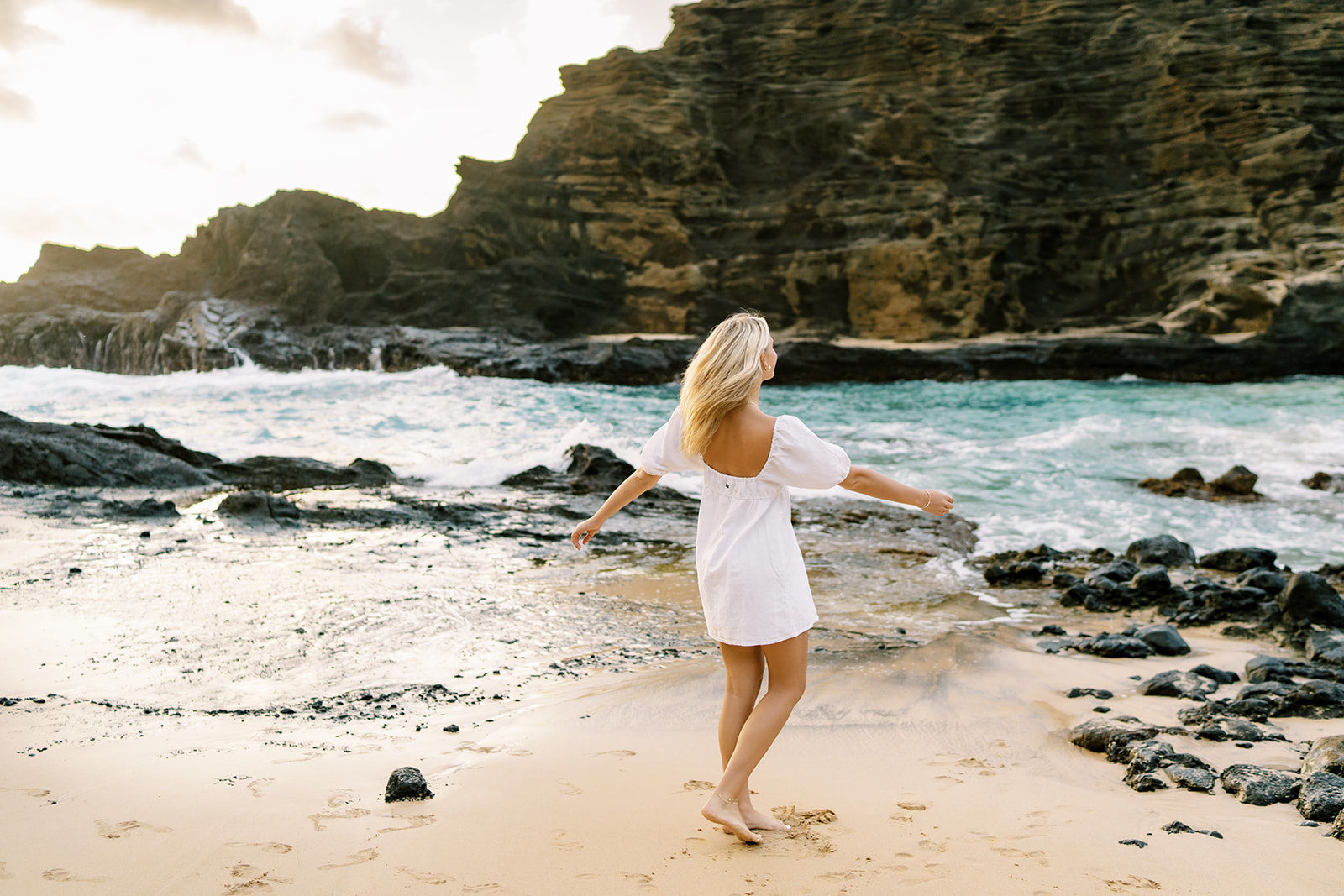 Woman in white dress walking on beach with arms outstretched, facing the ocean and cliffs in Halona Cove