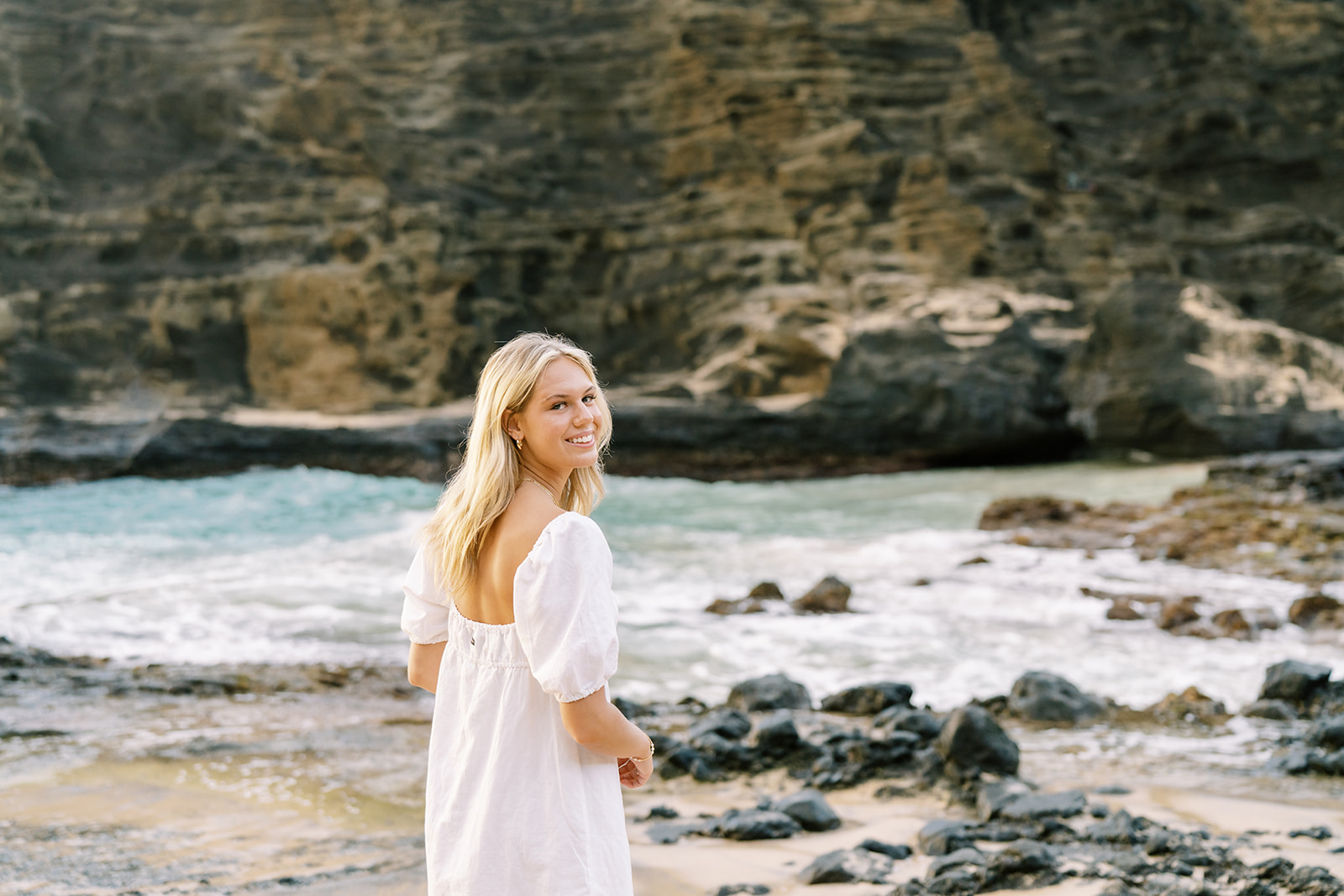 Woman in white dress standing on a beach looking over her shoulder with cliffs in front of her, Sunrise Portrait Session
