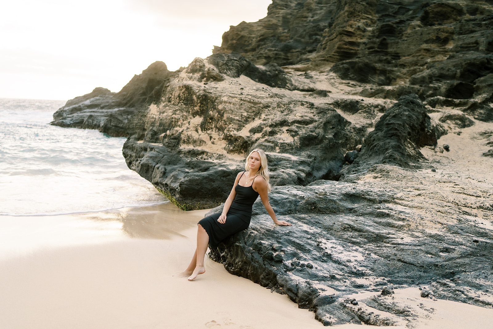 A woman in a black dress sitting on a rocky formation by the beach at Halona Cove