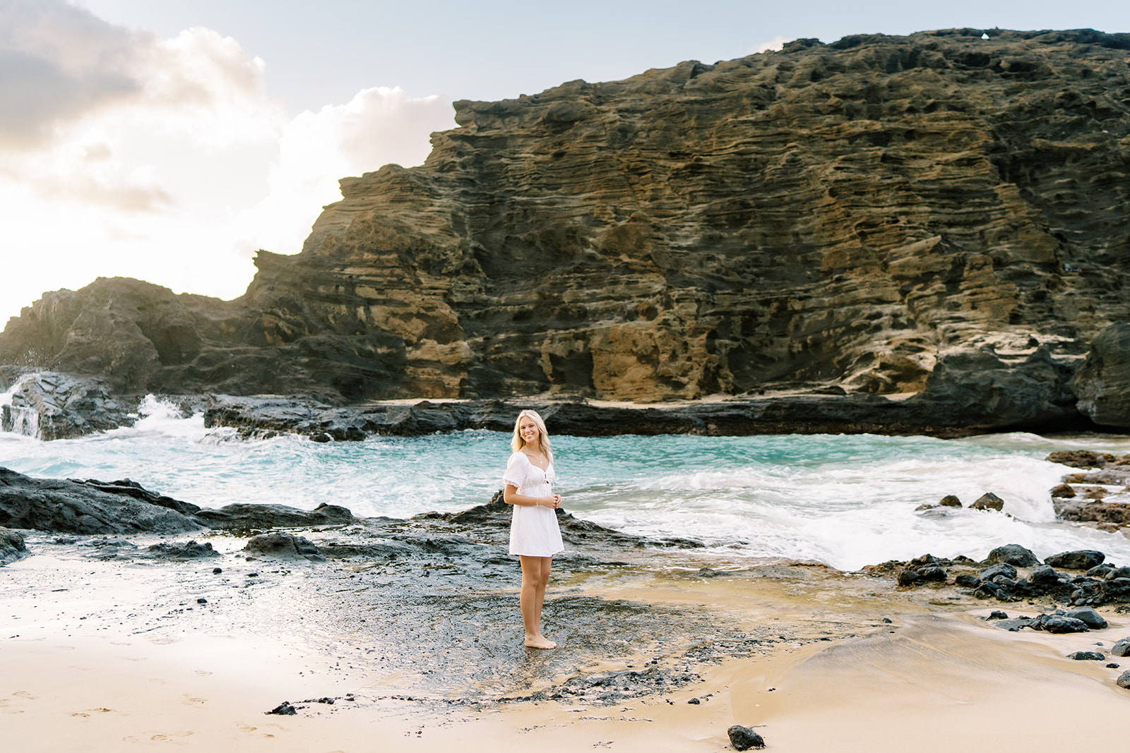 A woman in a white dress stands on a sandy beach with rocky cliffs and waves Portrait session at Halona Cove