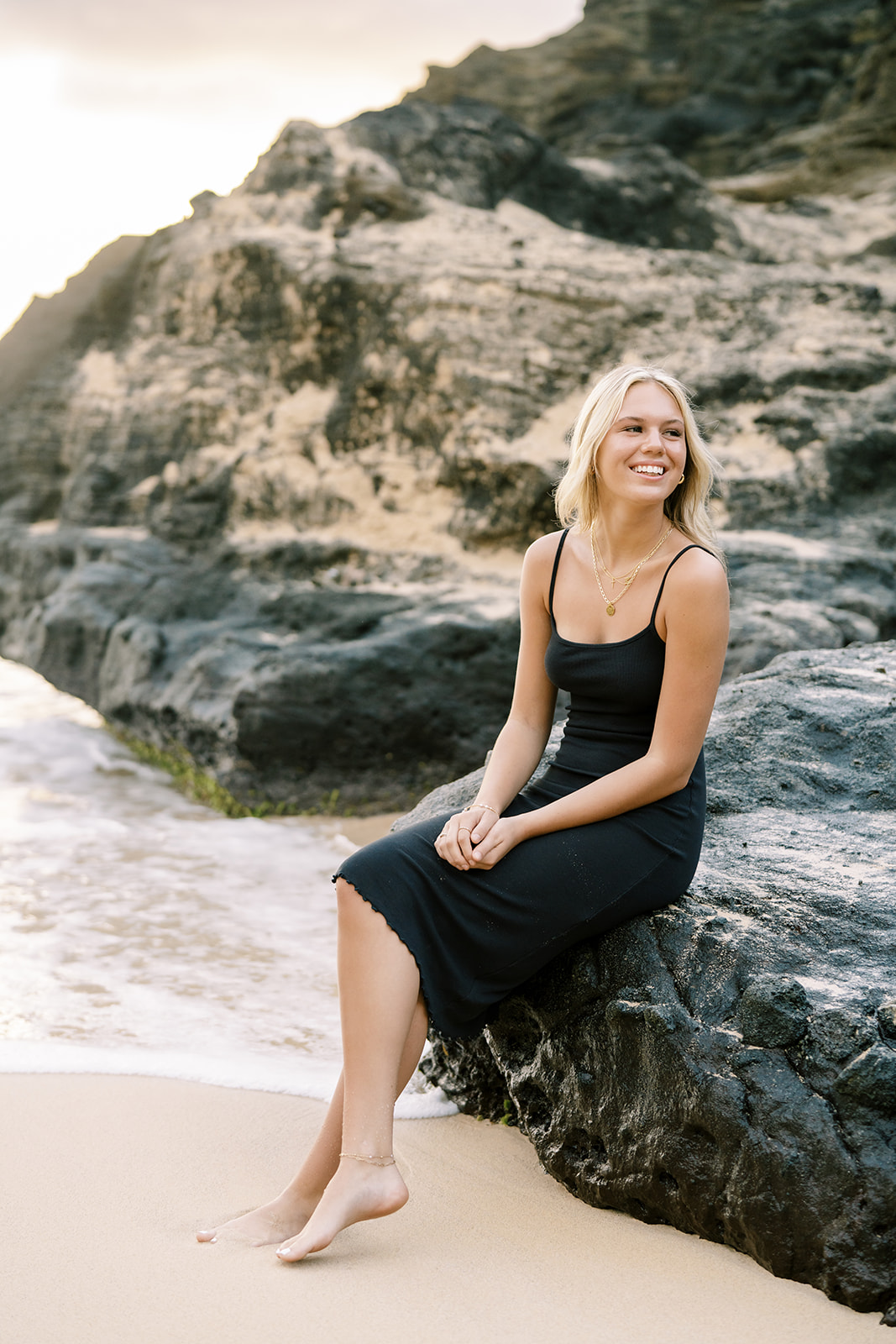 A smiling woman sitting on a rock at the beach with her feet in the sand during her high school senior portrait in Oahu