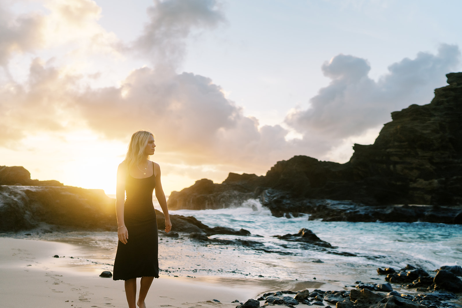 Woman standing on a beach at sunrise, with waves crashing on the shore and rocky cliffs senior portraits that go beyond 