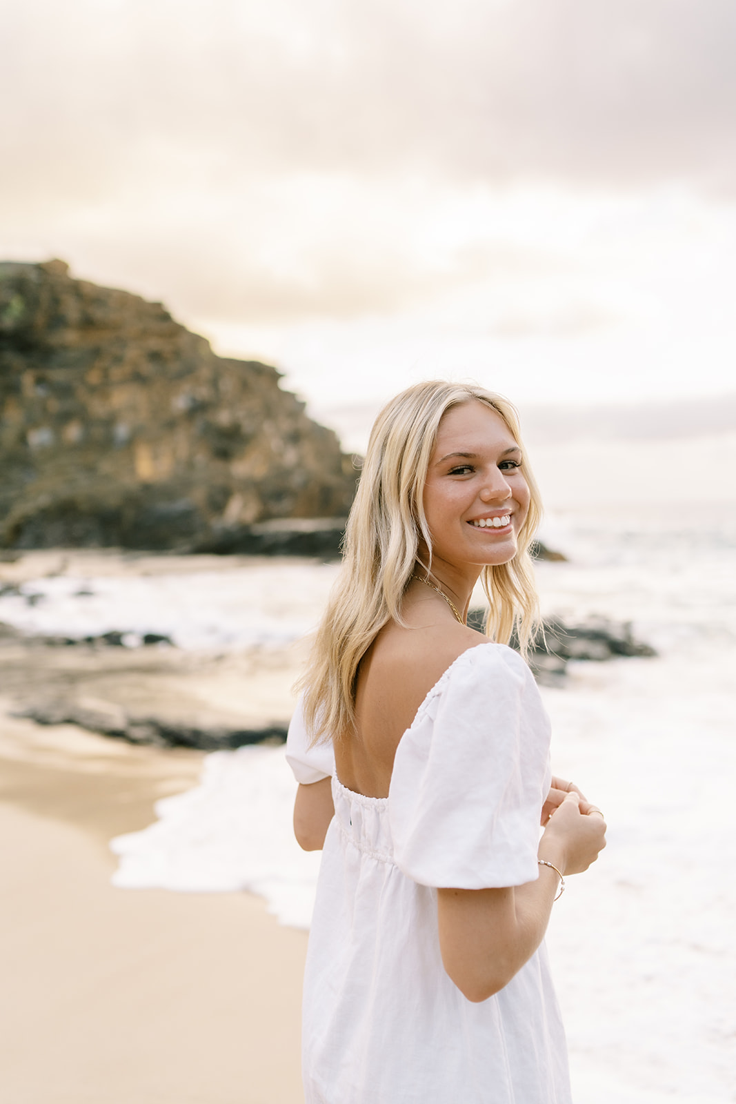 A smiling woman in a white dress standing on a beach with cliffs and the ocean in the background.