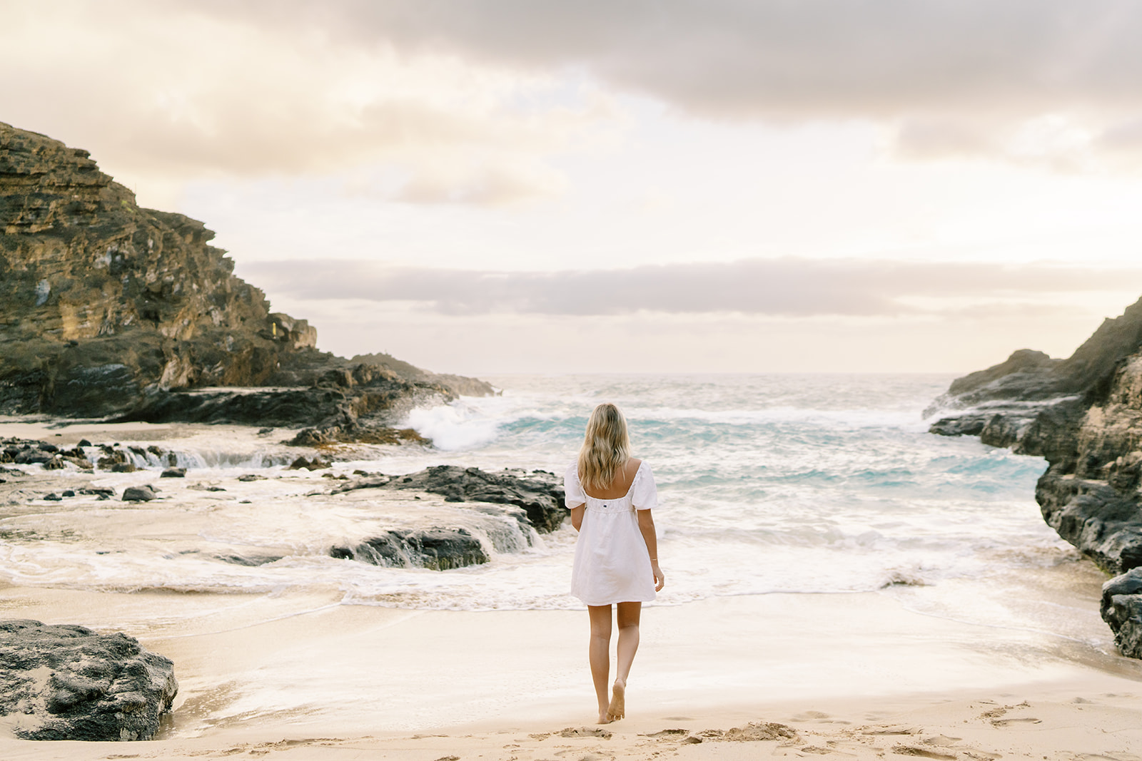 A woman in a white dress walking towards the ocean on a sandy beach with rocky cliffs on the side, Sunrise Portrait