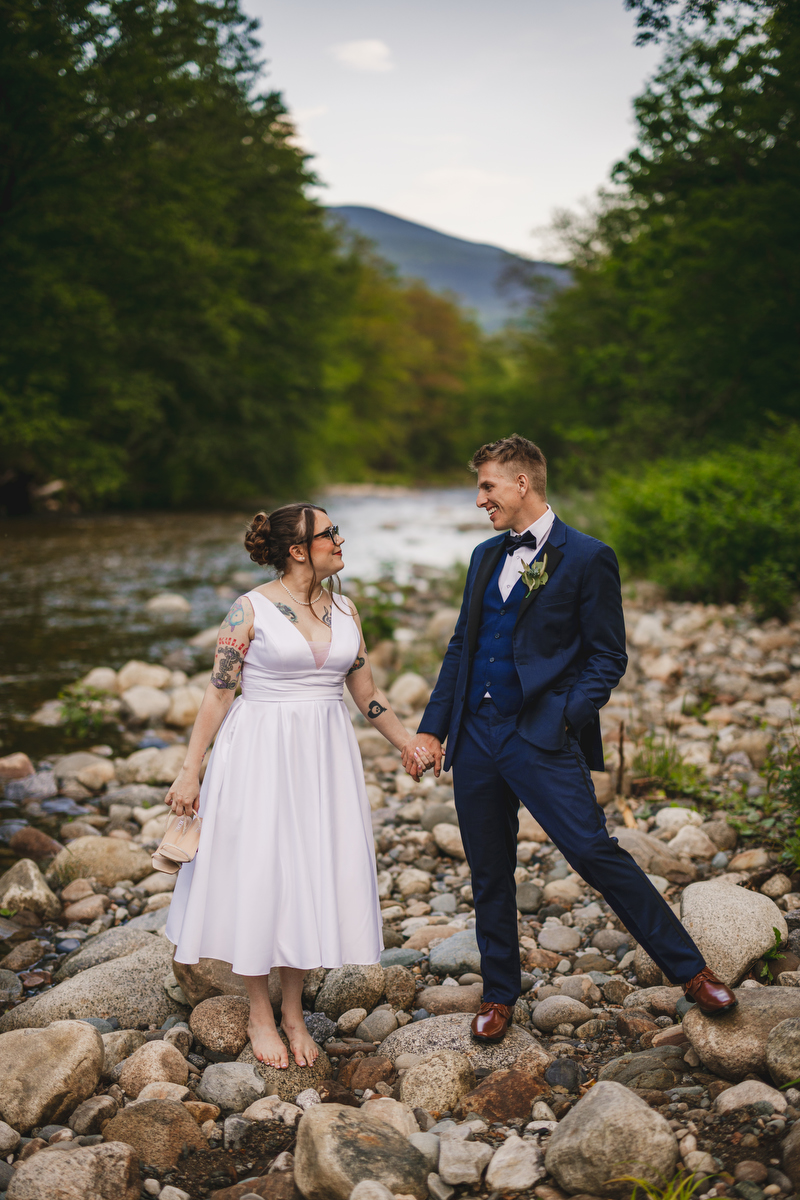 bride and groom stand on the rocks at the edge of a river