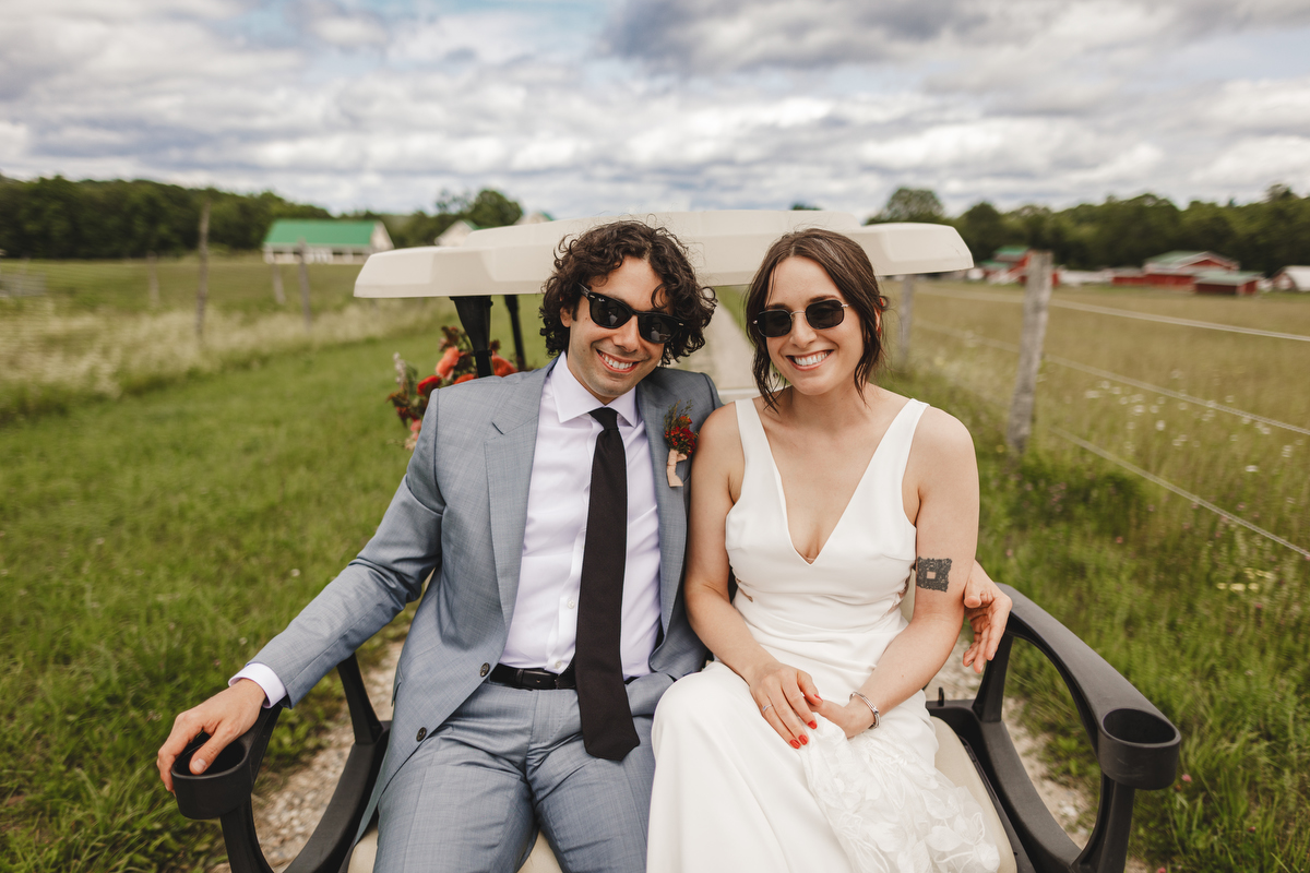 A bride and groom wearing sunglasses ride in a golf cart at Valley View Farm