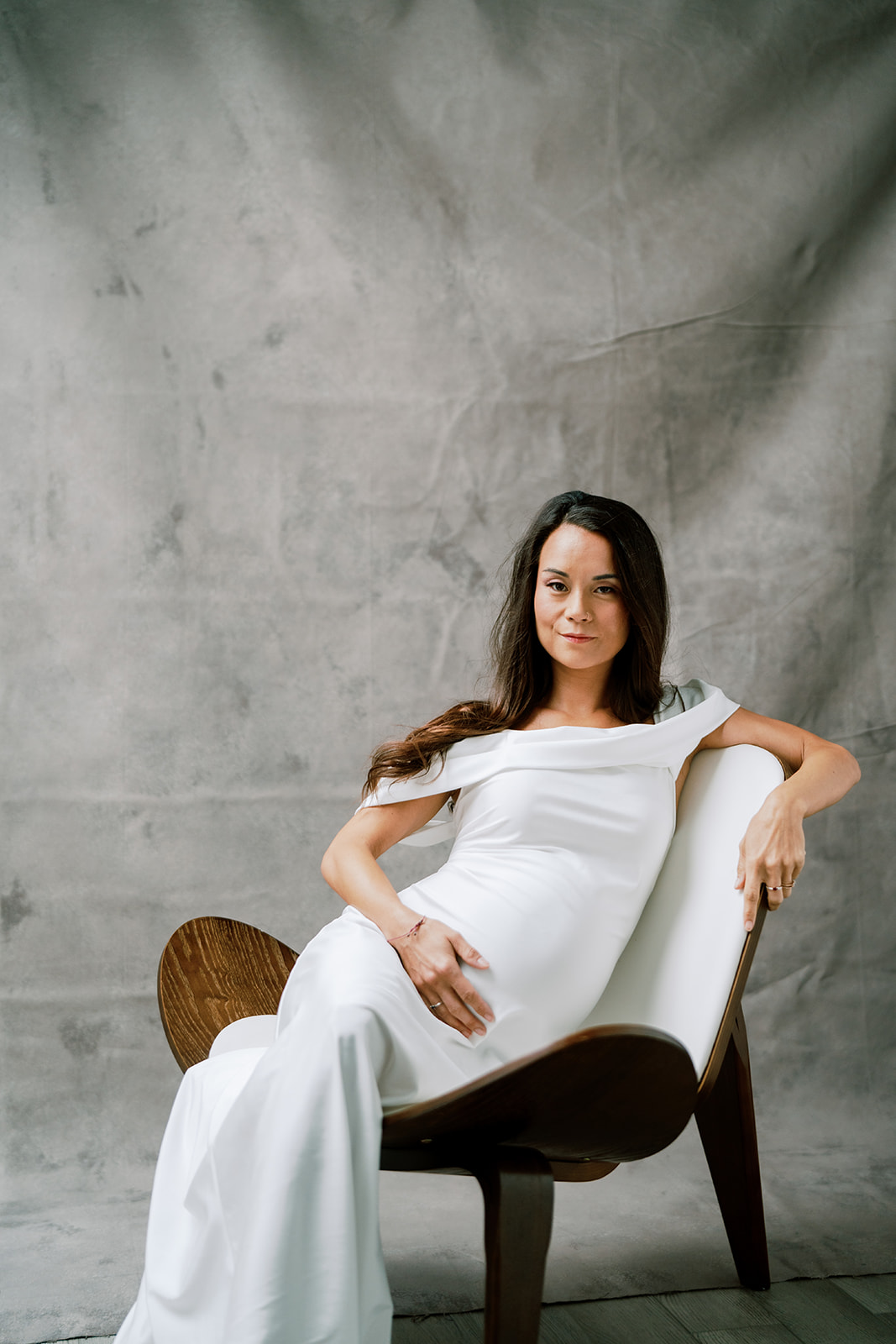 A pregnant woman in a white dress sitting in a chair looking straight to the camera.