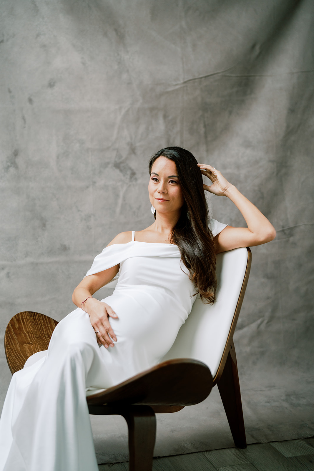 A pregnant woman in a white dress sitting in a chair during her maternity photography studio session.