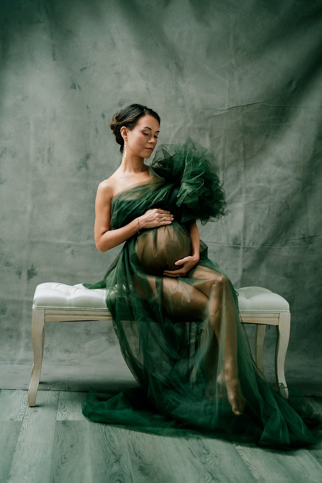 A pregnant woman in a green dress sitting on a bench during her studio maternity session.