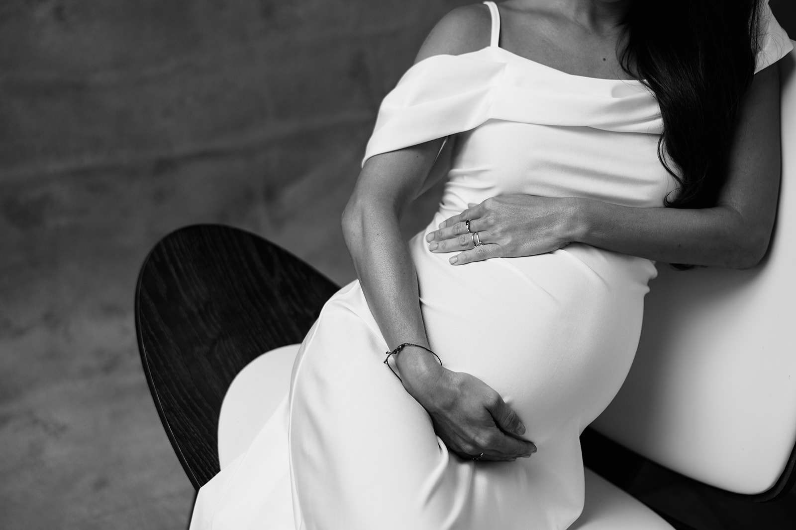 A monochrome photo of a pregnant woman in a white dress sitting on a chair during her portrait photography