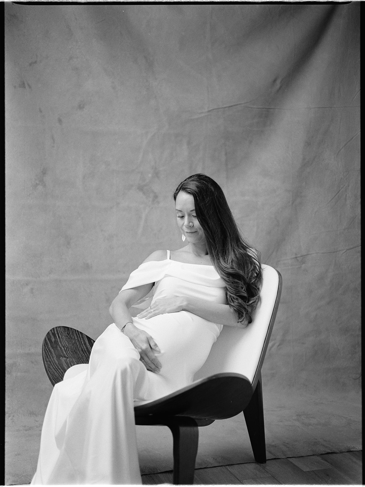 A pregnant woman in a white dress holding her baby bump while sitting in a chair.