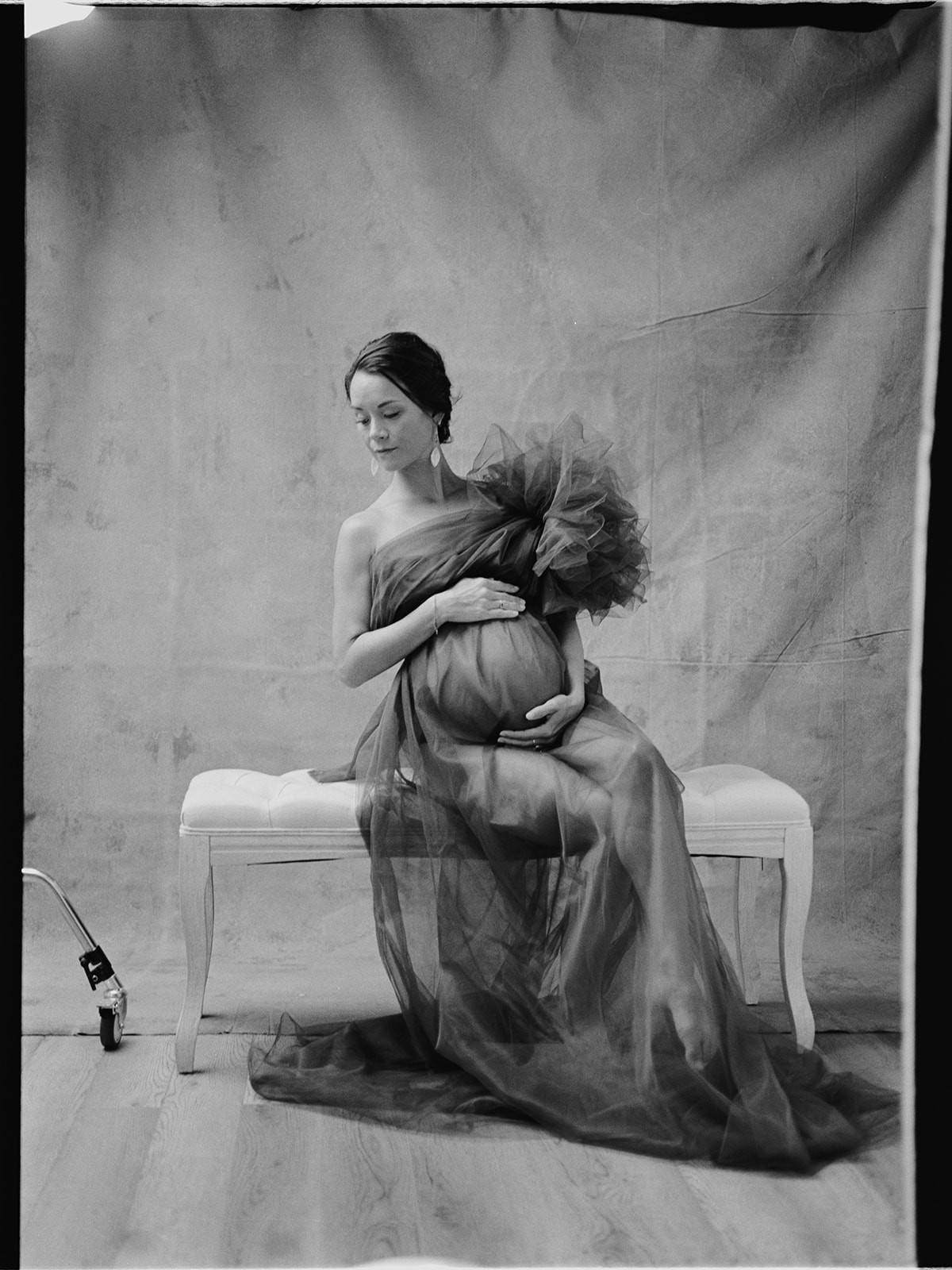 A black and white photo of a pregnant woman is sitting on a bench during her studio maternity session.
