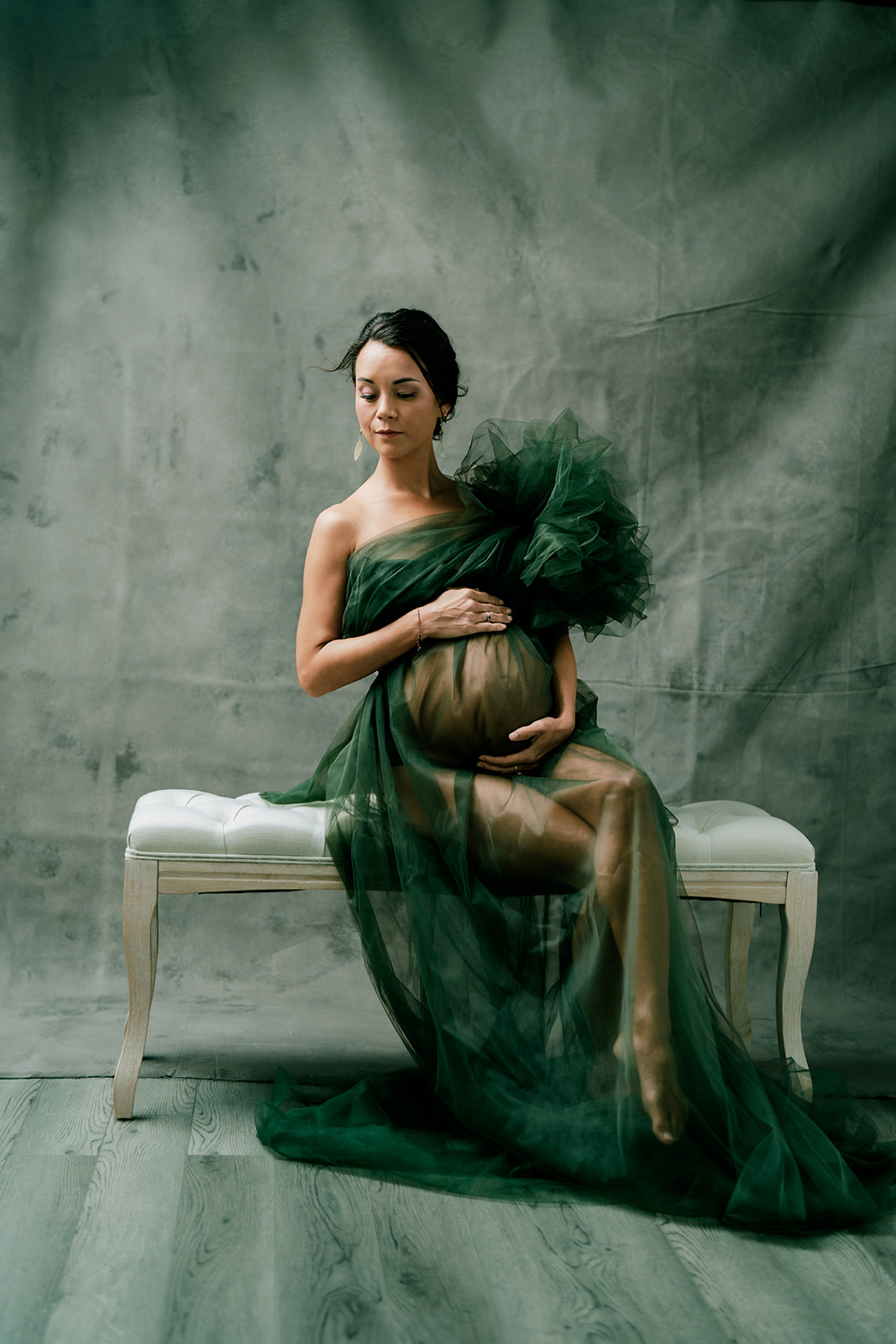 A pregnant woman wearing a green dress, sitting on a bench in studio.