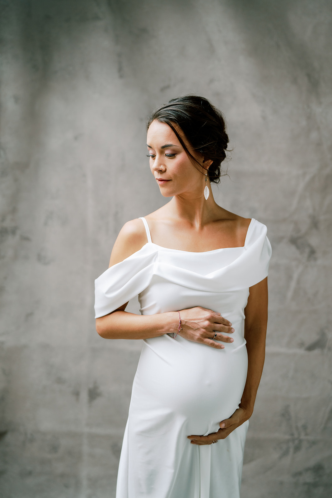 A pregnant woman holding her baby bump in a white dress posing for a photo. 