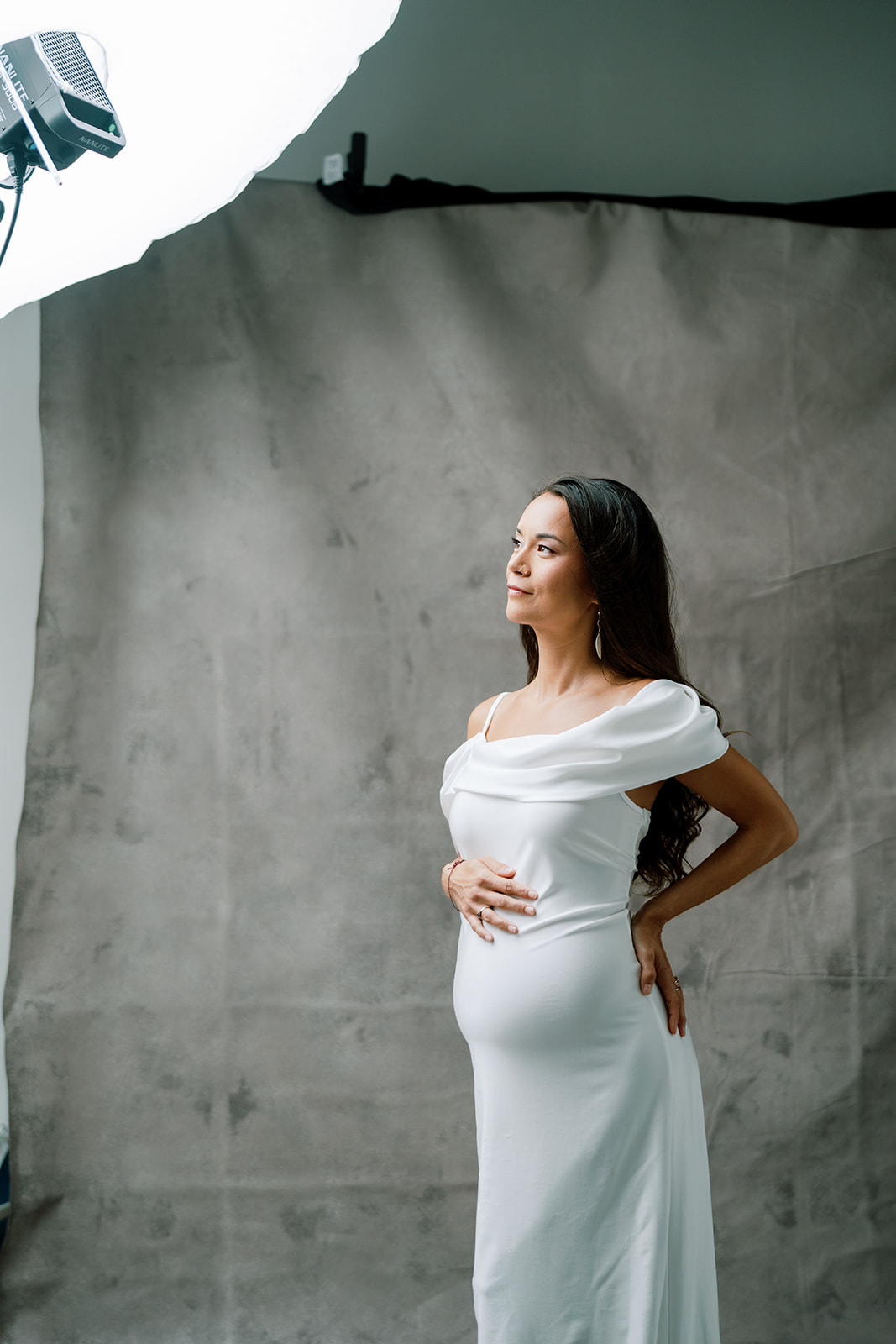 A pregnant woman in a white dress posing in front of a light during her studio maternity session.
