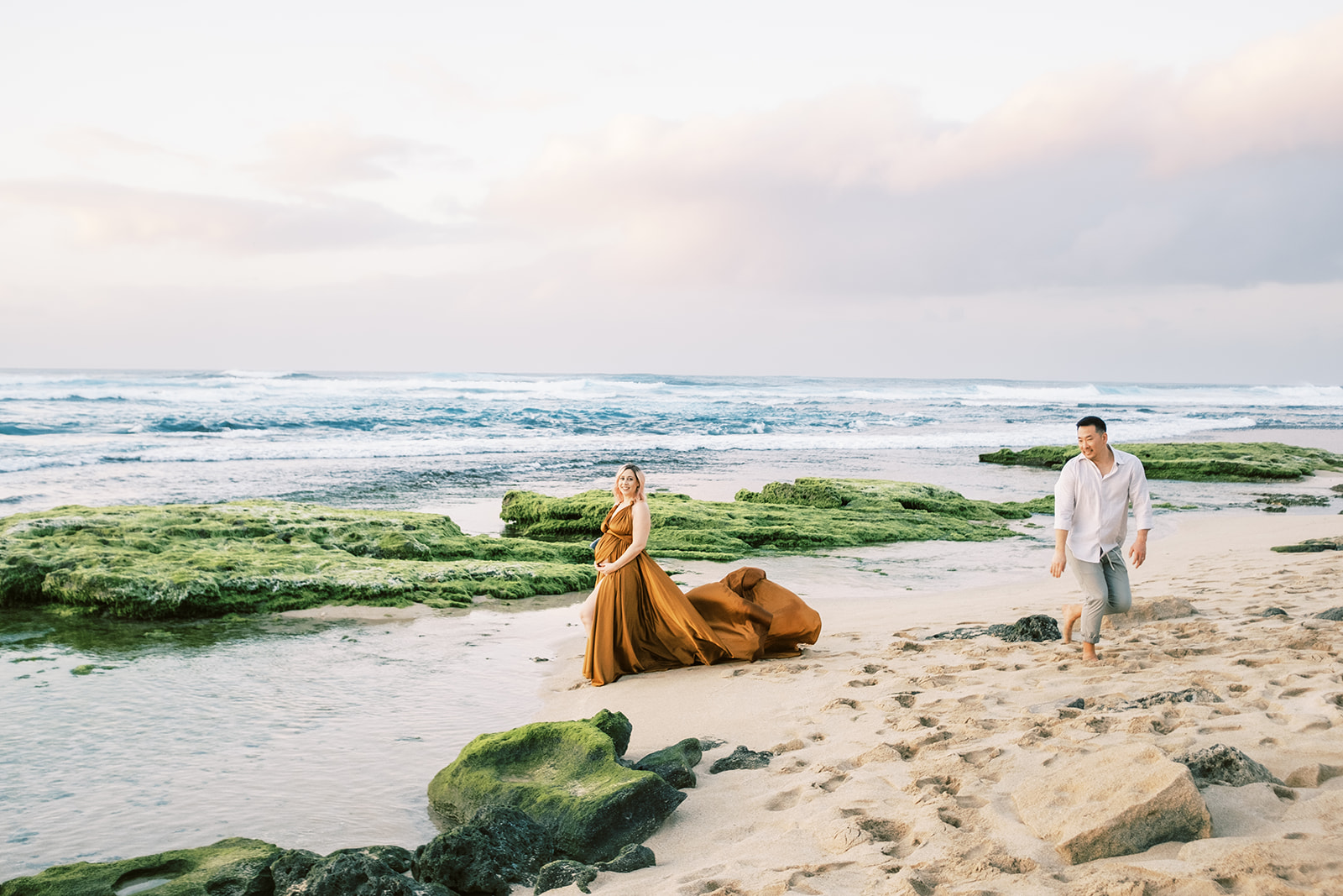 A couple walks on a beach with moss-covered rocks on Oahu during sunset Maternity Session with Megan Moura