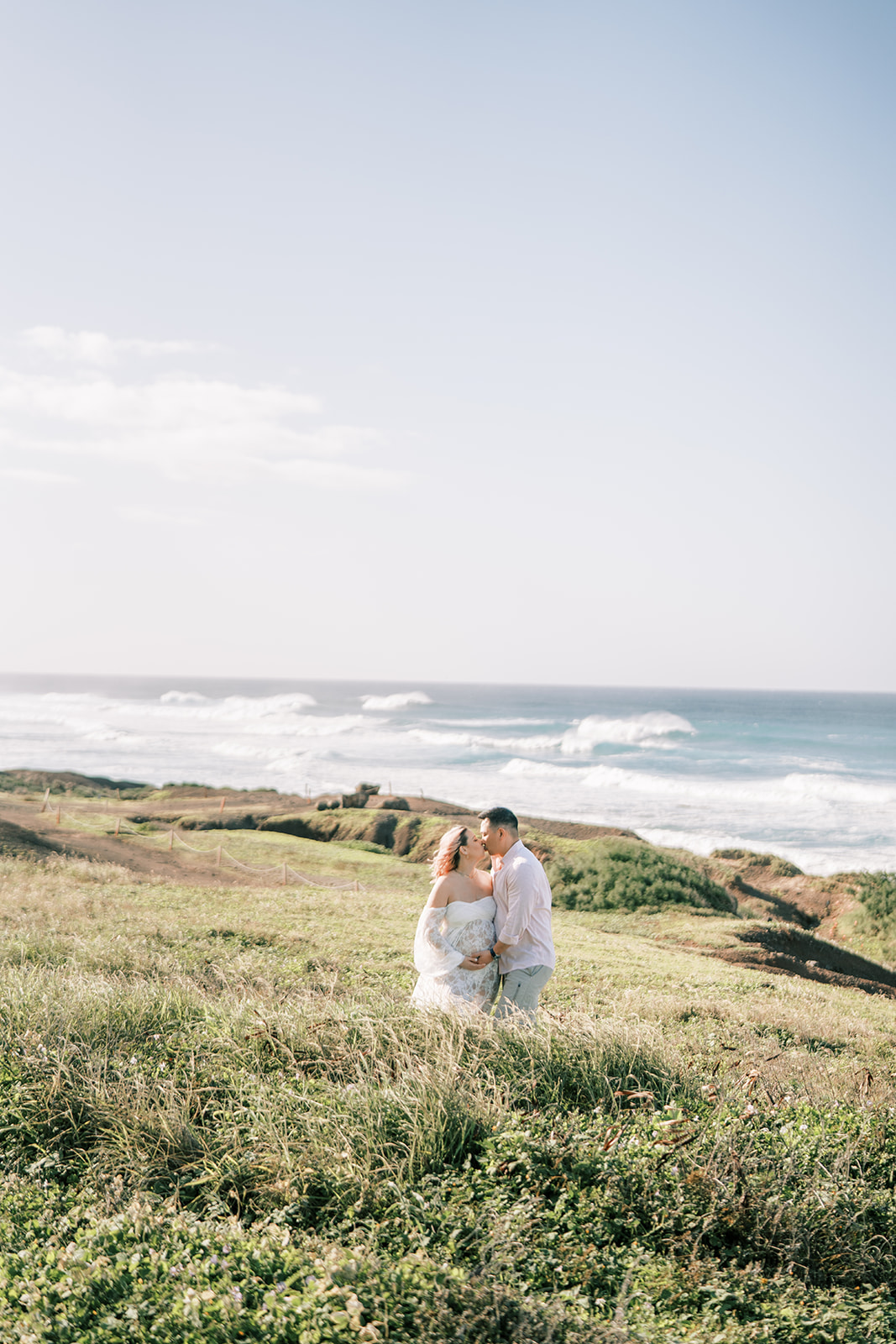 A couple embracing in a coastal grassy field with the ocean in the background during their Maternity Session