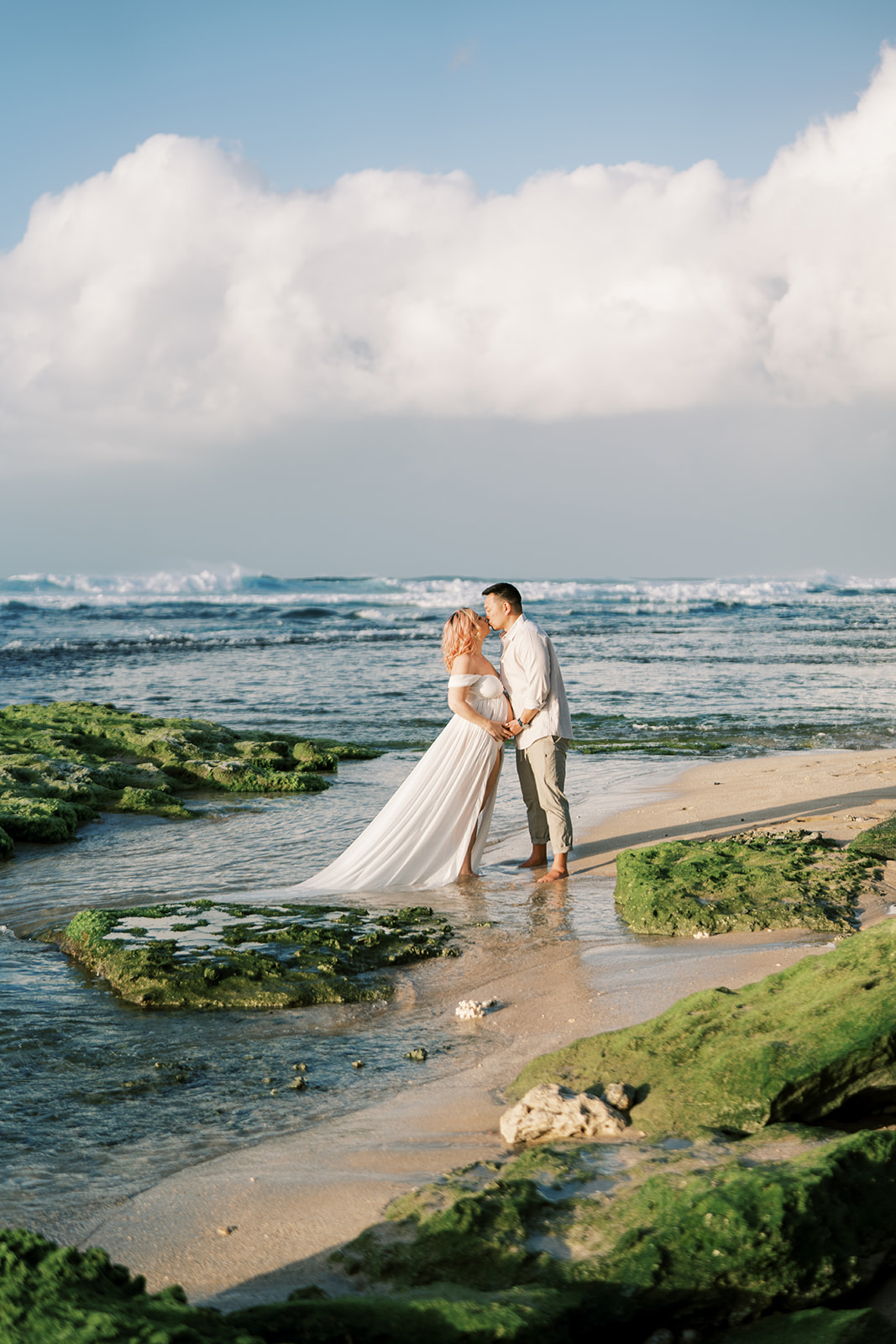 Couple in wedding attire kissing on a beach with green moss-covered rocks Maternity session captured by Megan Moura Oahu