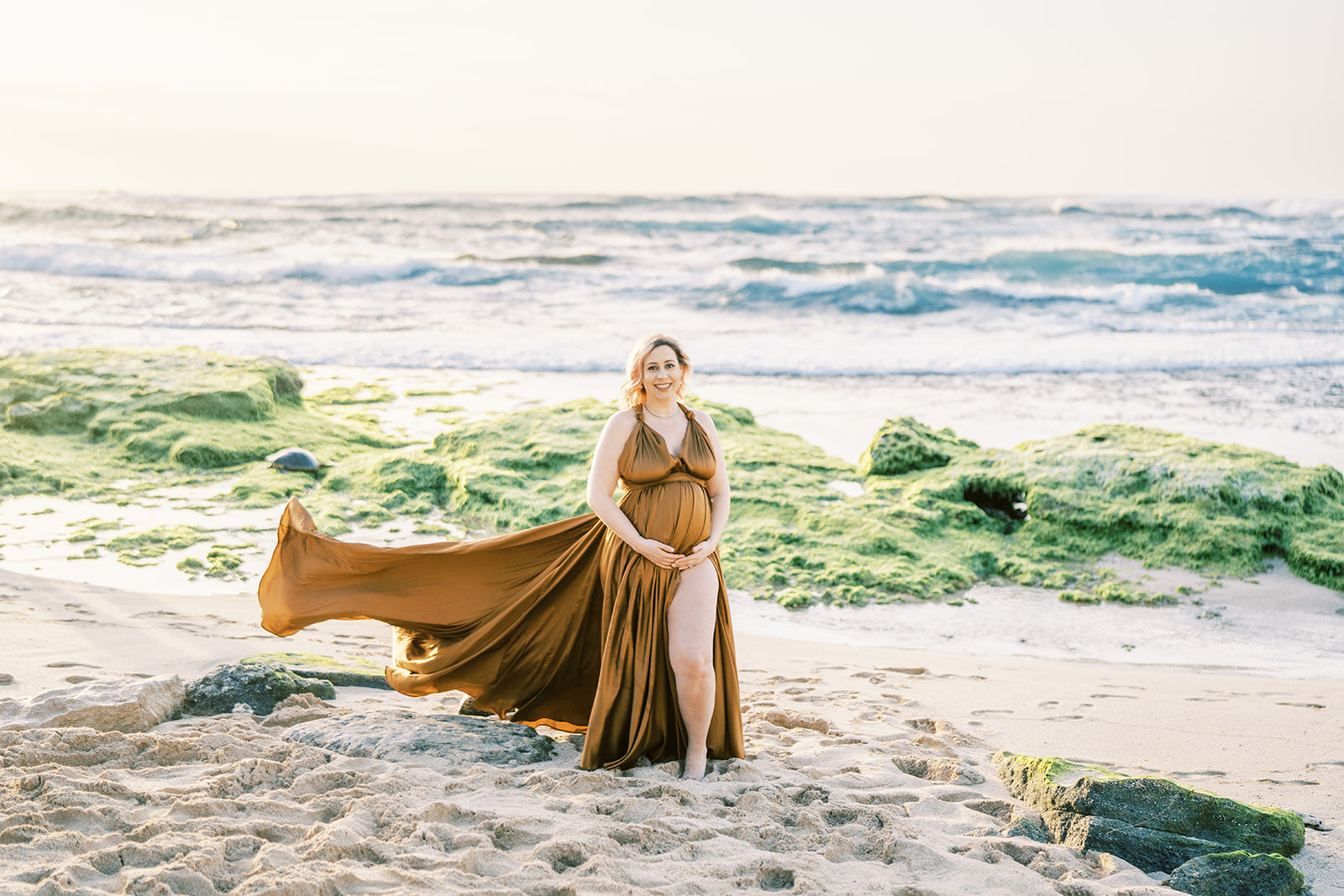 Pregnant woman in a flowing dress posing on the beach with the ocean in the background during sunset Maternity Session