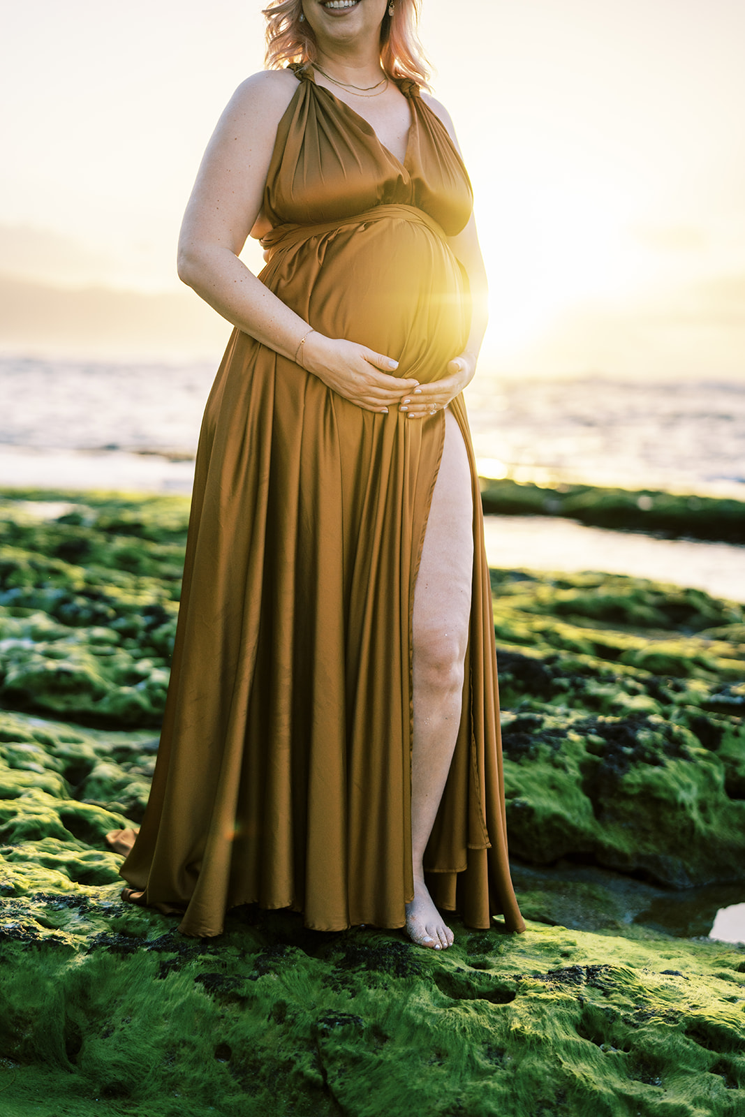 Pregnant woman standing on a rocky beach at sunset, cradling her belly during sunset Maternity Session on Oahu