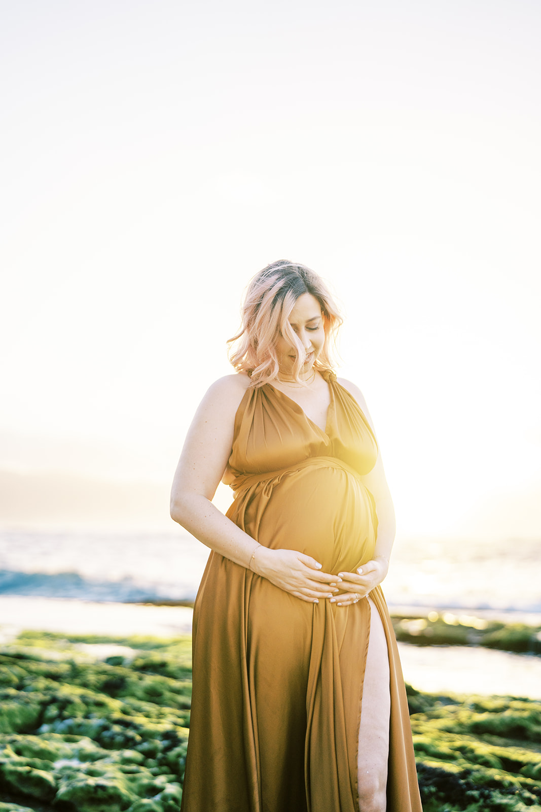 Pregnant woman in a yellow dress holding her baby bump and looking down during sunset Maternity Session on Oahu