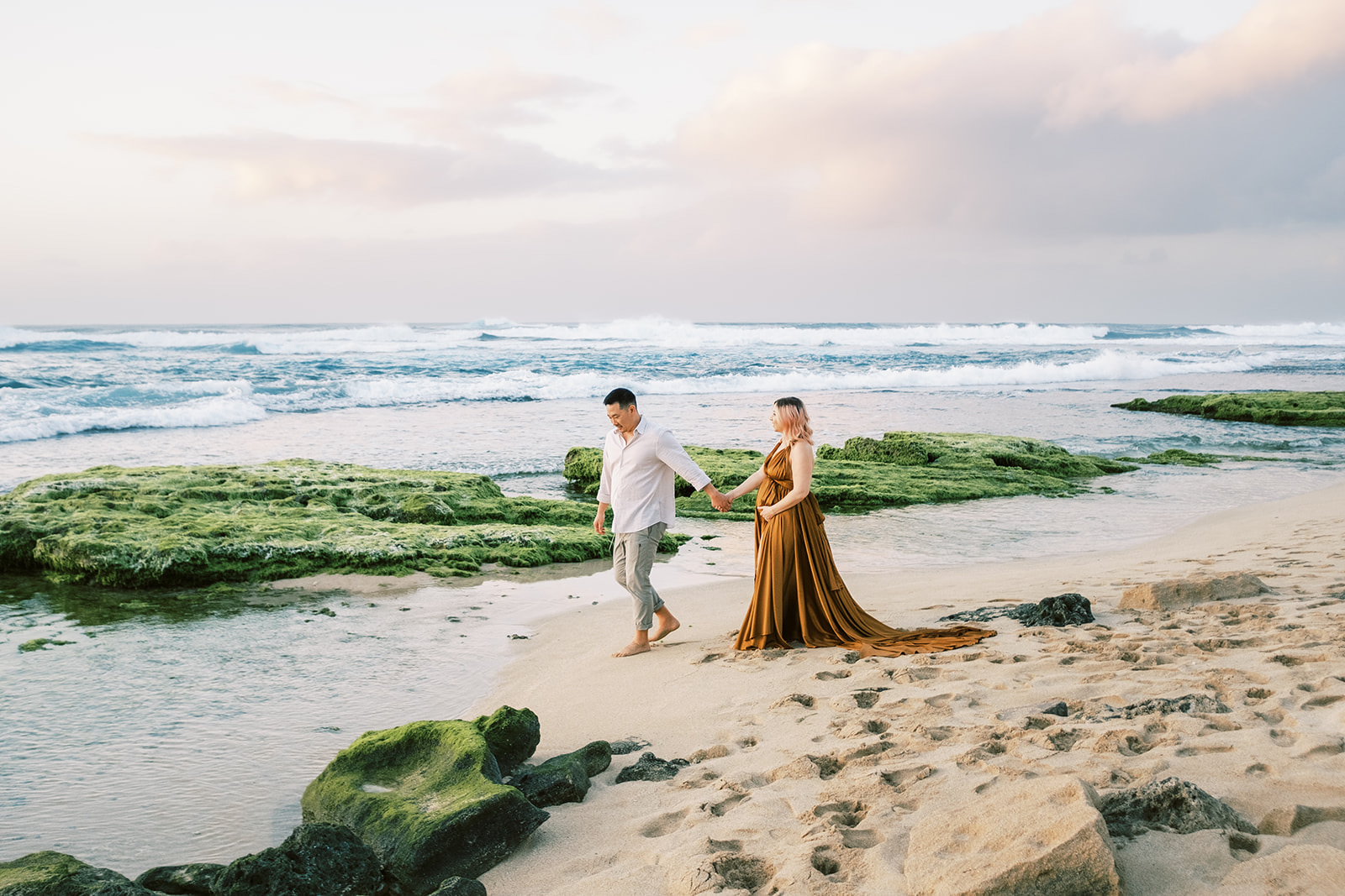 Couple holding hands during their Maternity Session on Oahu, walking on a beach with mossy rocks and ocean waves.