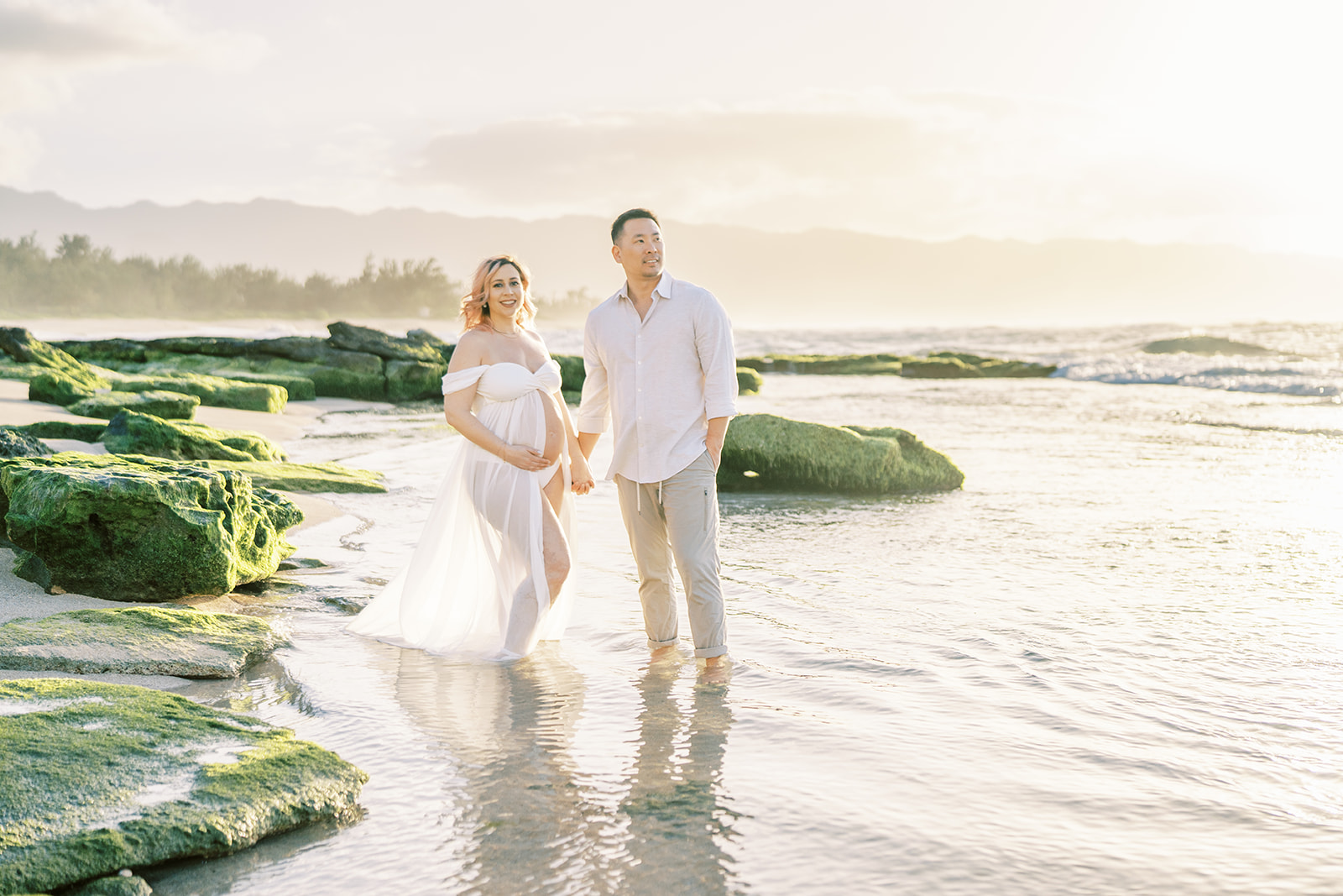 A couple expecting a child holding hands and walking on a beach at sunset on Oahu