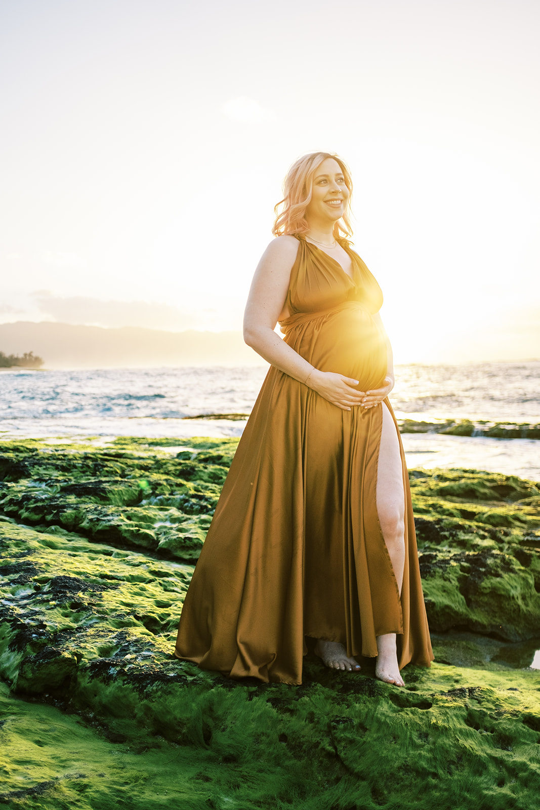 Pregnant woman in a flowing dress standing on a moss-covered shore during sunset Maternity Session on Oahu