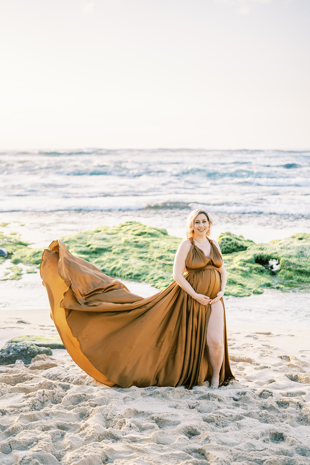 A pregnant woman smiling at the camera with her brown dress flowing in the breeze sunset Maternity Session on Oahu