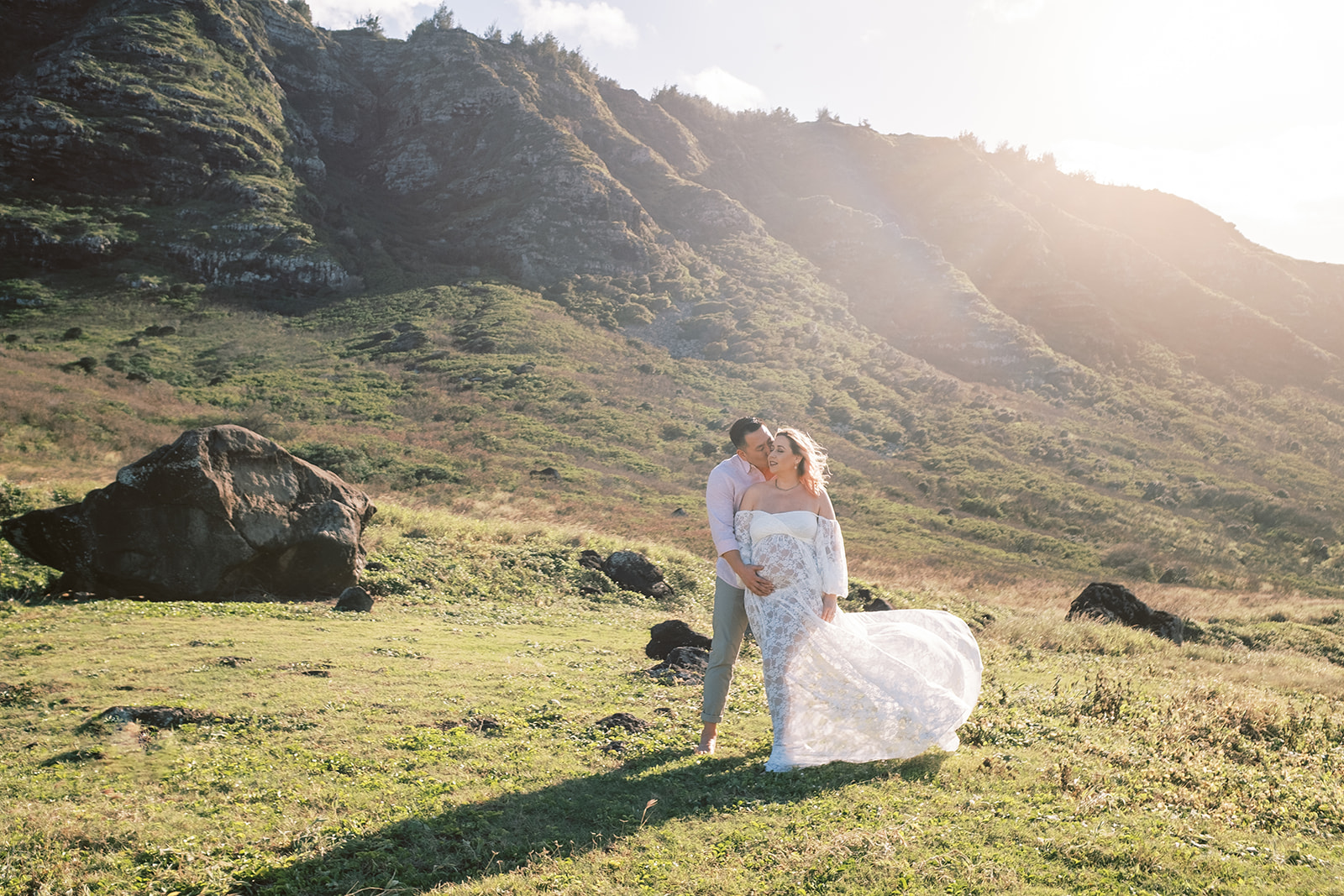 A couple in wedding attire sharing a kiss in a sunlit field with mountains, captured by an Oahu photographer.