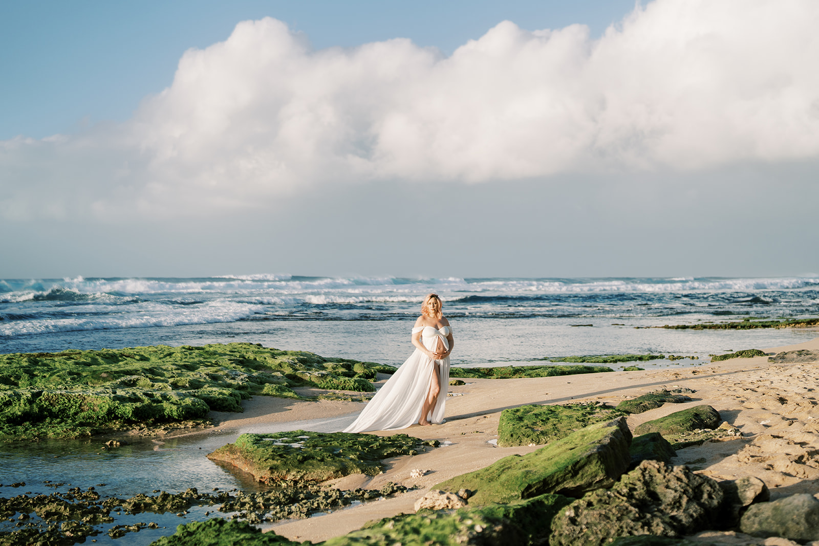 A pregnant woman in a white dress standing on a beach with waves in the background Maternity session captured by Megan M