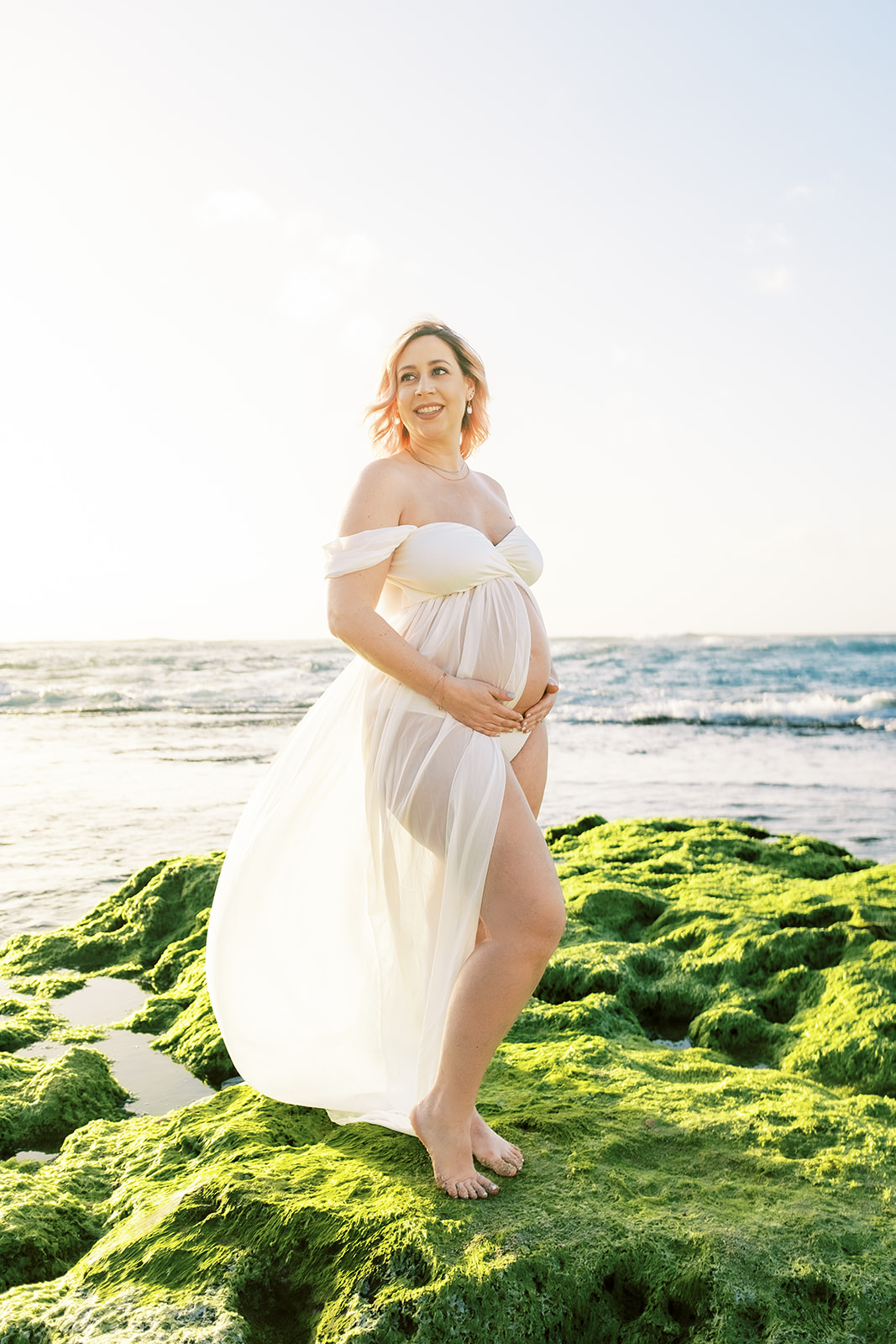 A pregnant woman in a white dress holding her baby bump on a beach with waves in the background Maternity session