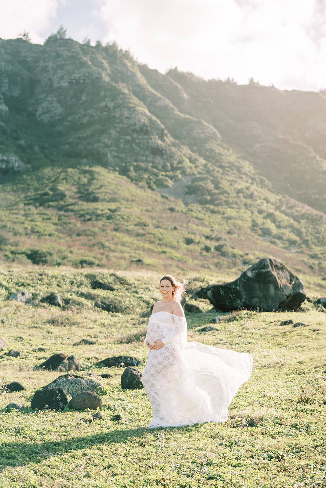 A pregnant woman in white dress twirling in a sunny, grassy field with hills in the background, by an Oahu photographer.