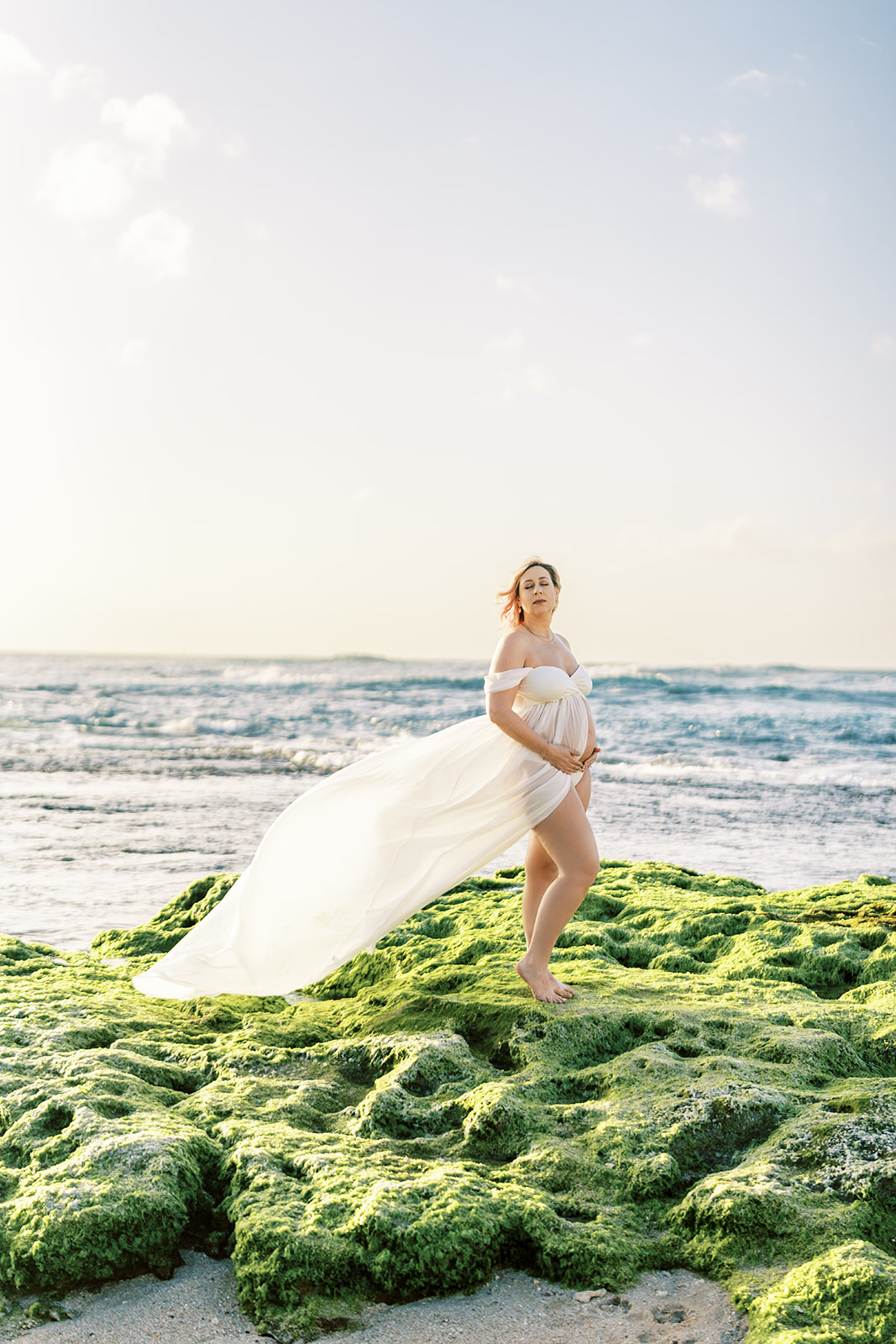 Pregnant woman in white flowing maternity outfit standing on a moss-covered rock by the sea during sunset.