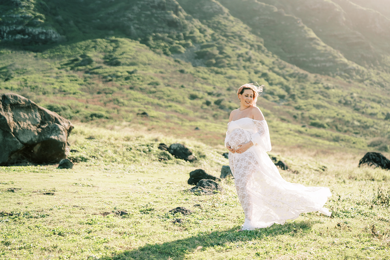 A pregnant woman in a white lace dress holding her baby bump in a sunny, grassy field with hills.