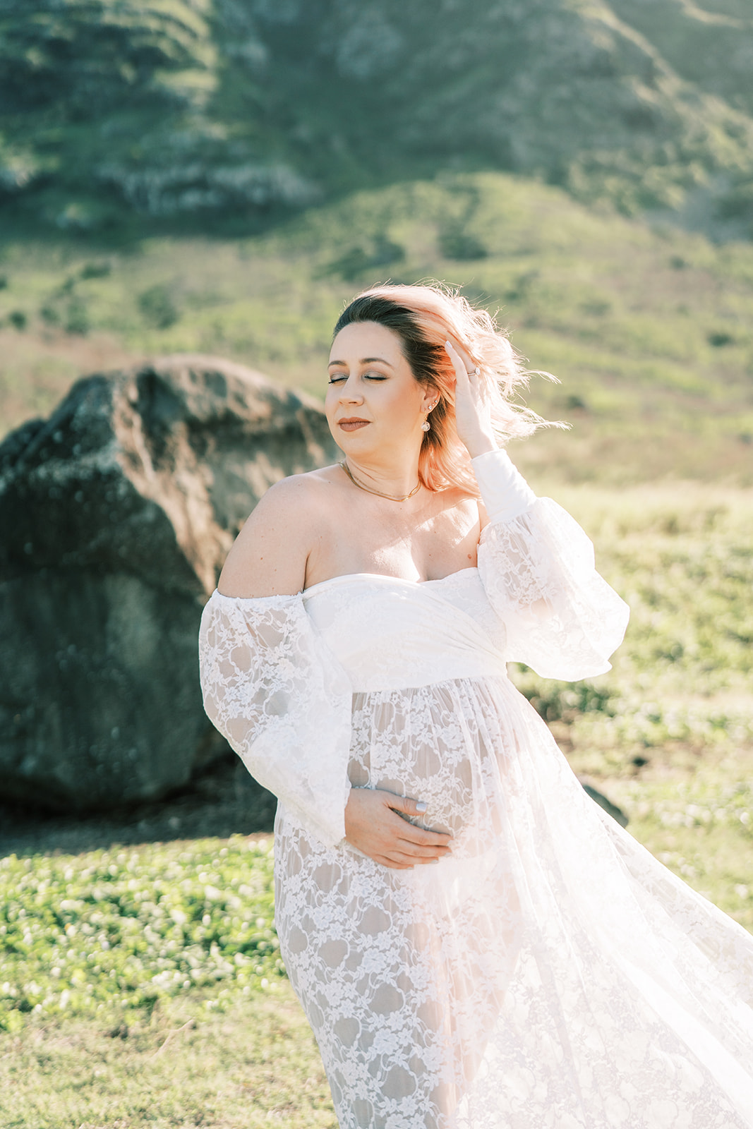 A pregnant woman in a white lace dress holding her hair back in a sunny, grassy field with a hill.