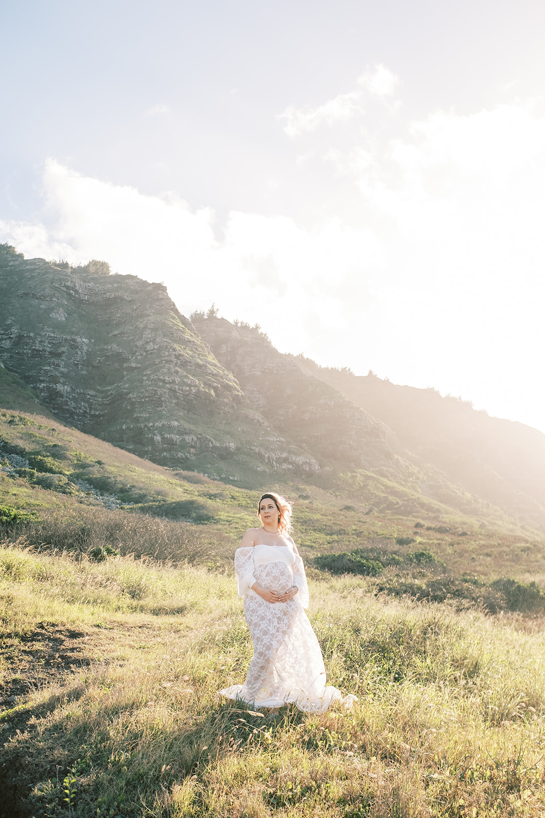 A pregnant woman in a white lace dress standing in a sunlit field, looking away with a mountain in the background