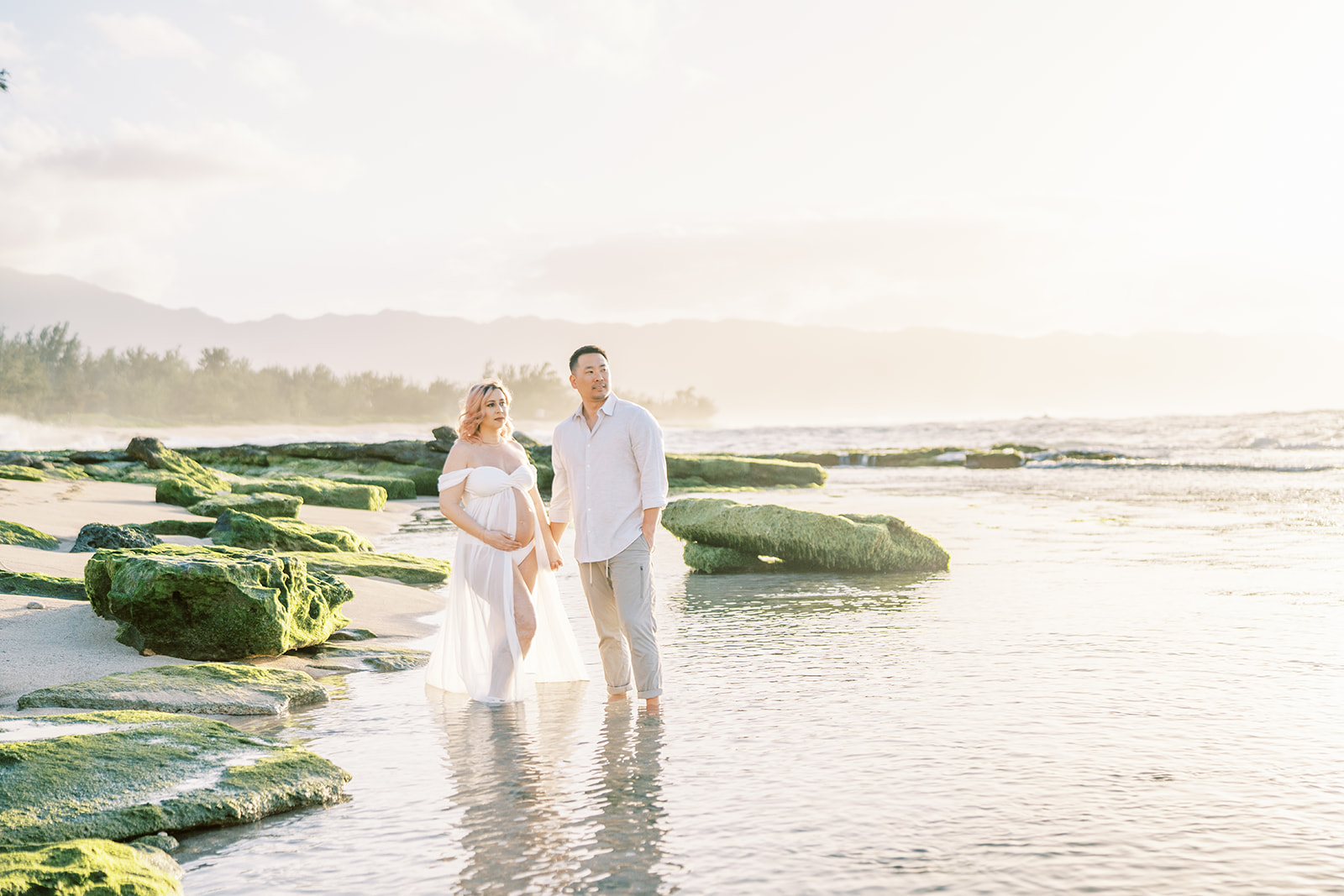 Couple wearing white clothing holding hands on a beach at sunset on Oahu Maternity Session.