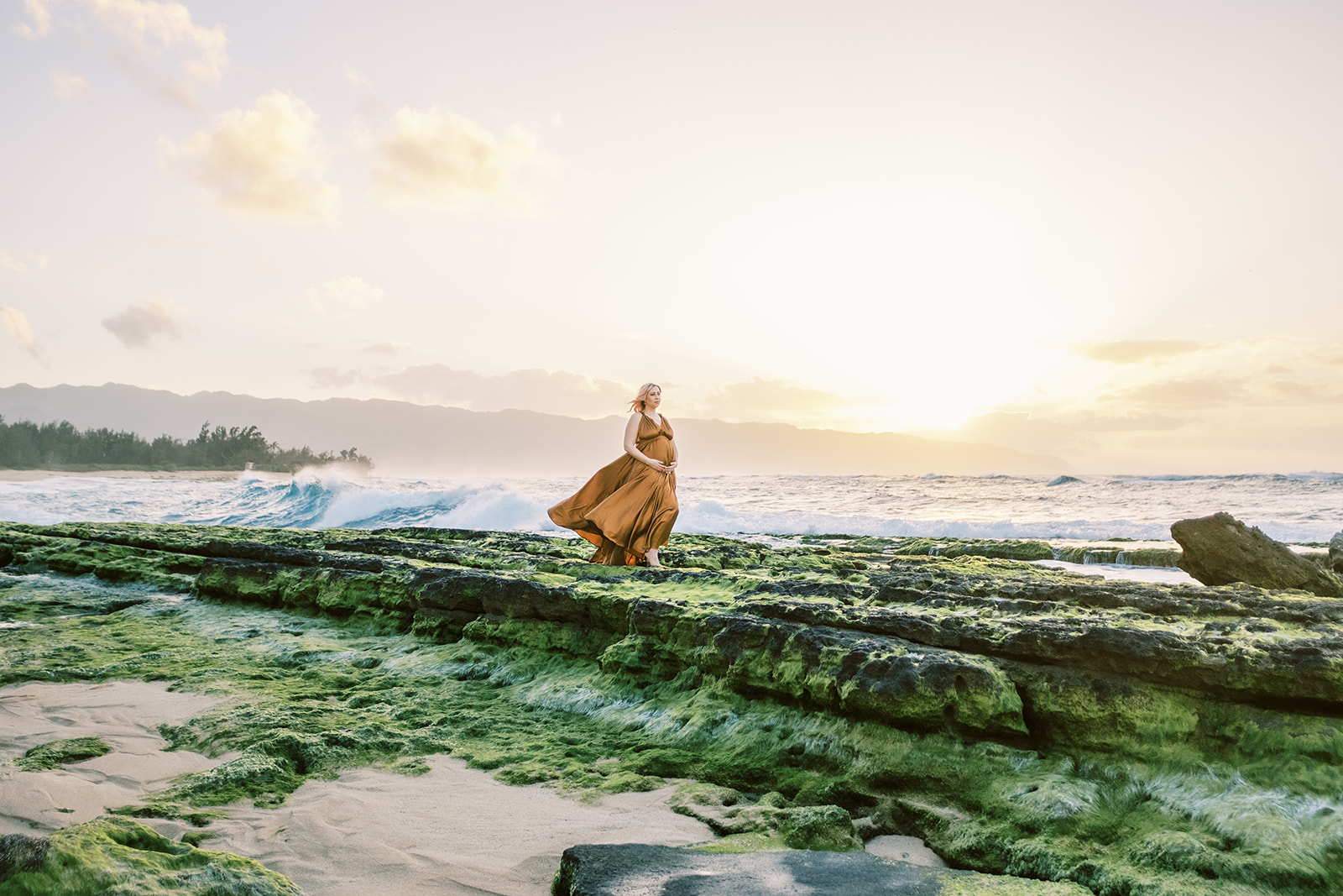 Woman in a flowing dress walking on a moss-covered rock by the sea at sunset during their maternity session on Oahu.