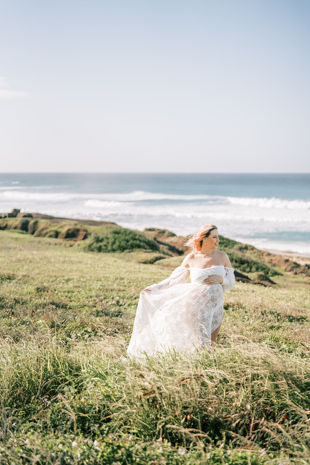 A woman in a white dress standing on a grassy field by the sea for her Maternity Session on Oahu.