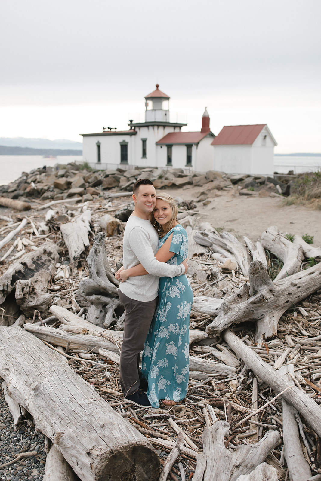 Engagement session in Seattle with couple standing on a log with the lighthouse visible in the background.