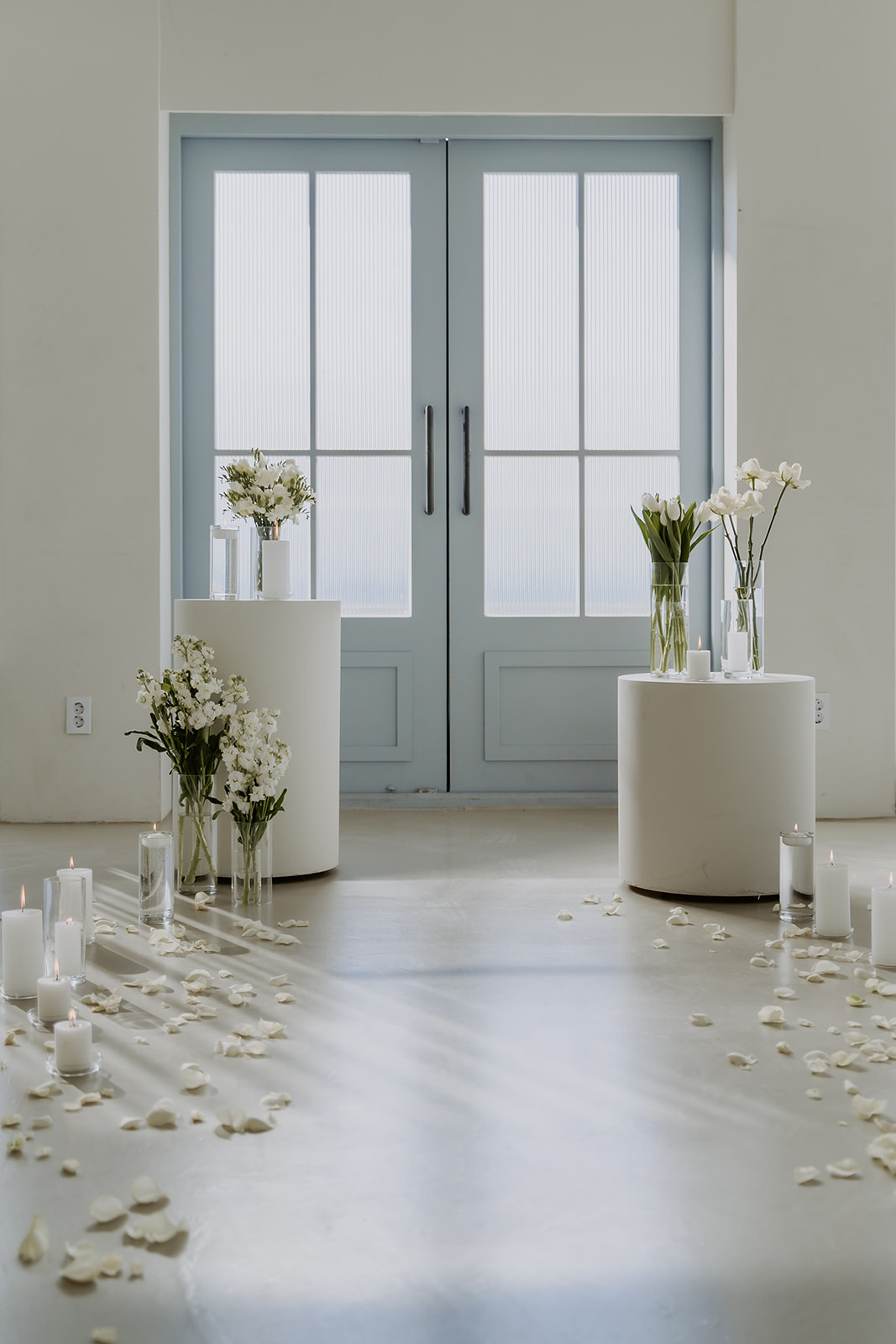 A serene room with a pair of french doors, white flowers in vases, scattered petals, and lit candles.