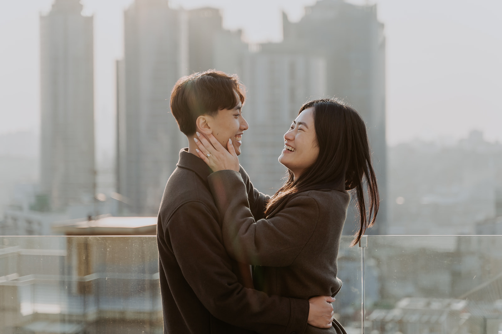 Two individuals share a tender moment on a rooftop with a Seoul cityscape background.