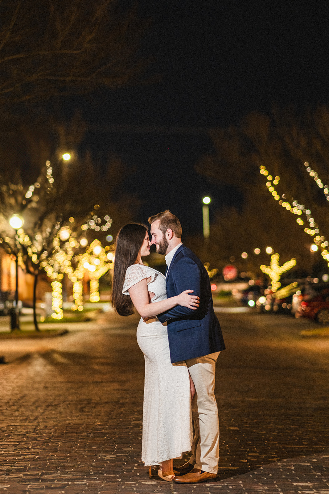 engaged couple embraces in the streets of downtown opelika alabama at night with the trees lit up behind them. 