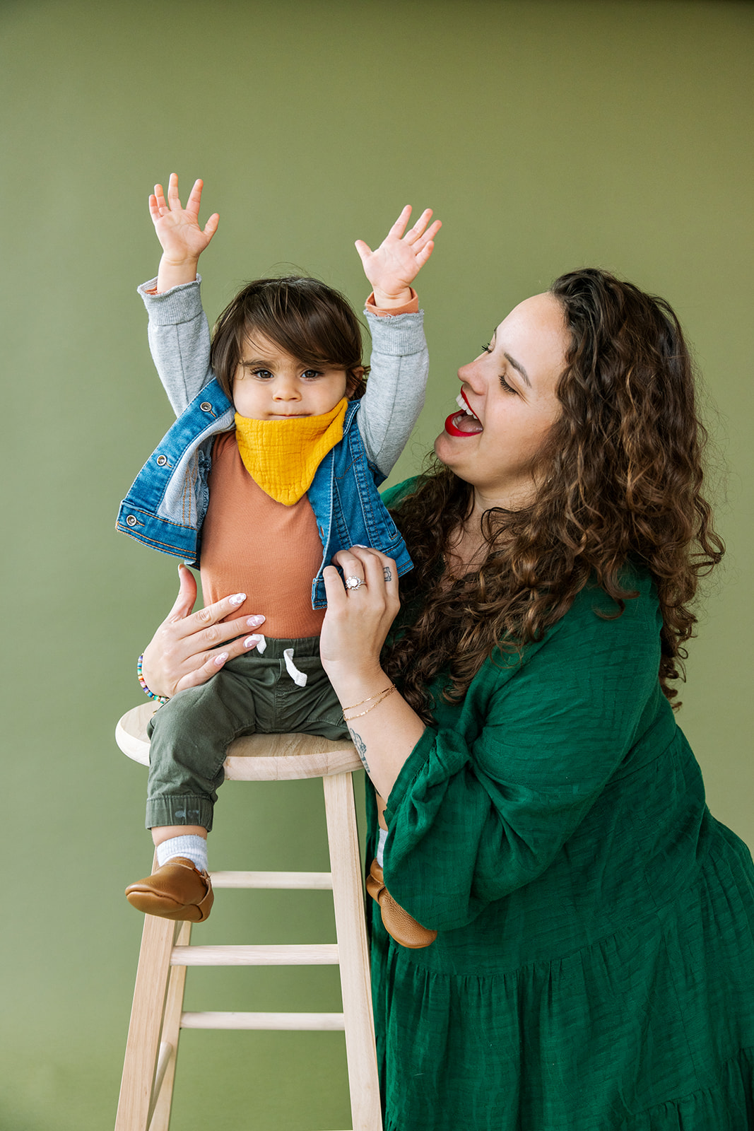 photo of toddler sitting on wooden stool raising his arms while mom holds onto him and steadies him